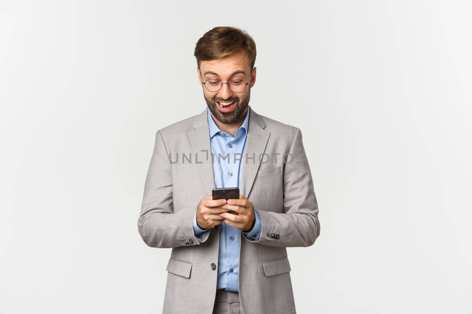 Portrait of handsome businessman with beard, wearing glasses and grey suit, looking surprised at mobile phone, reading something online, standing over white background by Benzoix