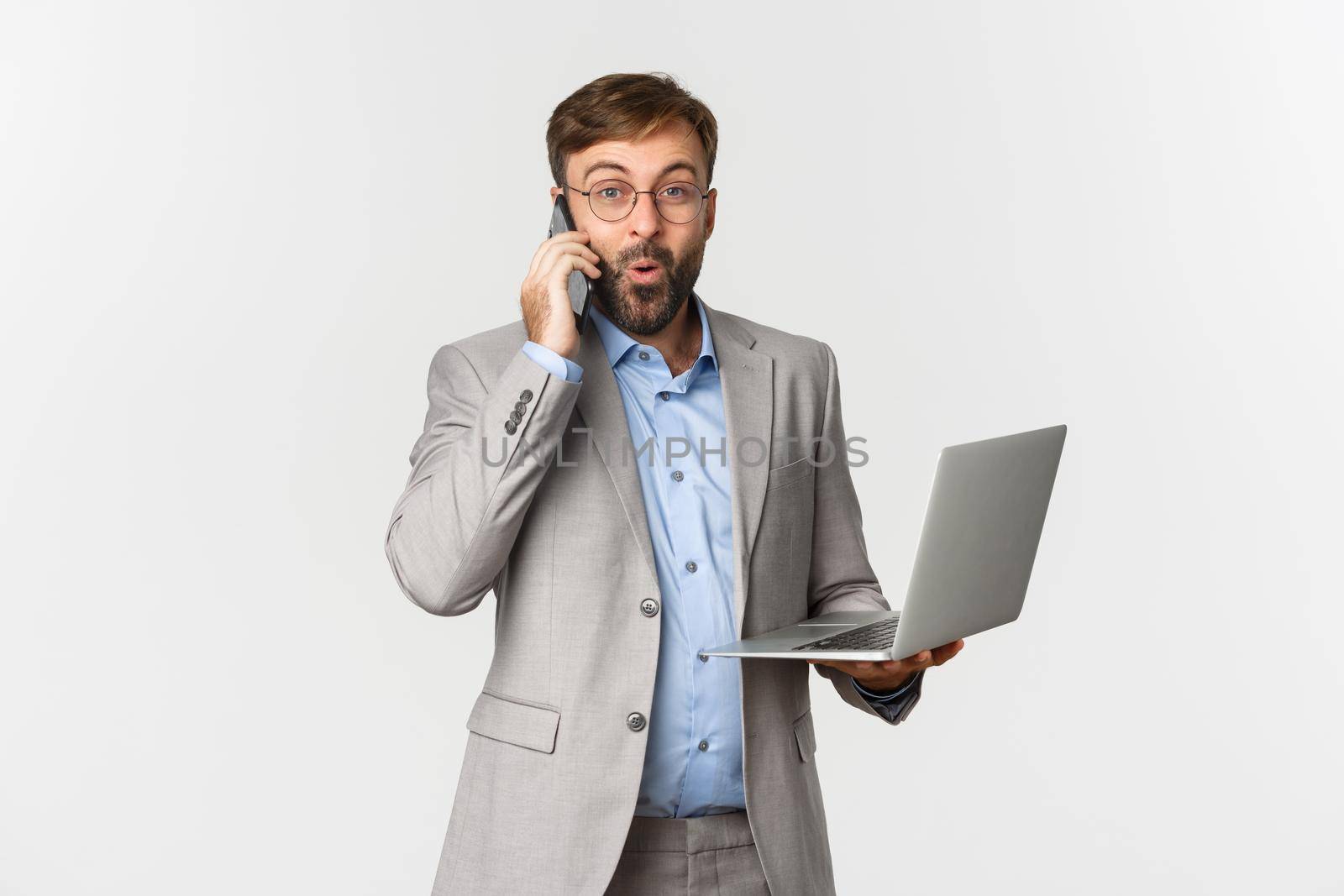 Portrait of handsome businessman in grey suit and glasses, receive good news during phone call, holding laptop, looking satisfied, standing over white background.