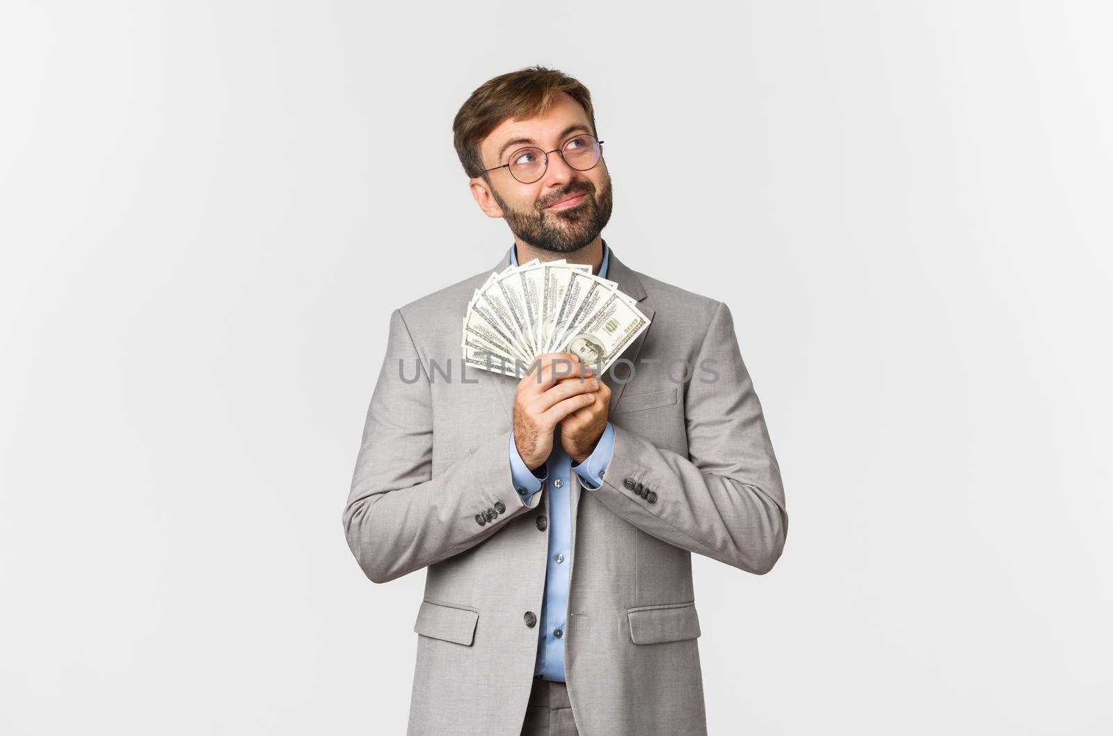 Image of happy businessman with beard, wearing grey suit and glasses, holding money and thinking about shopping or investing, standing over white background.