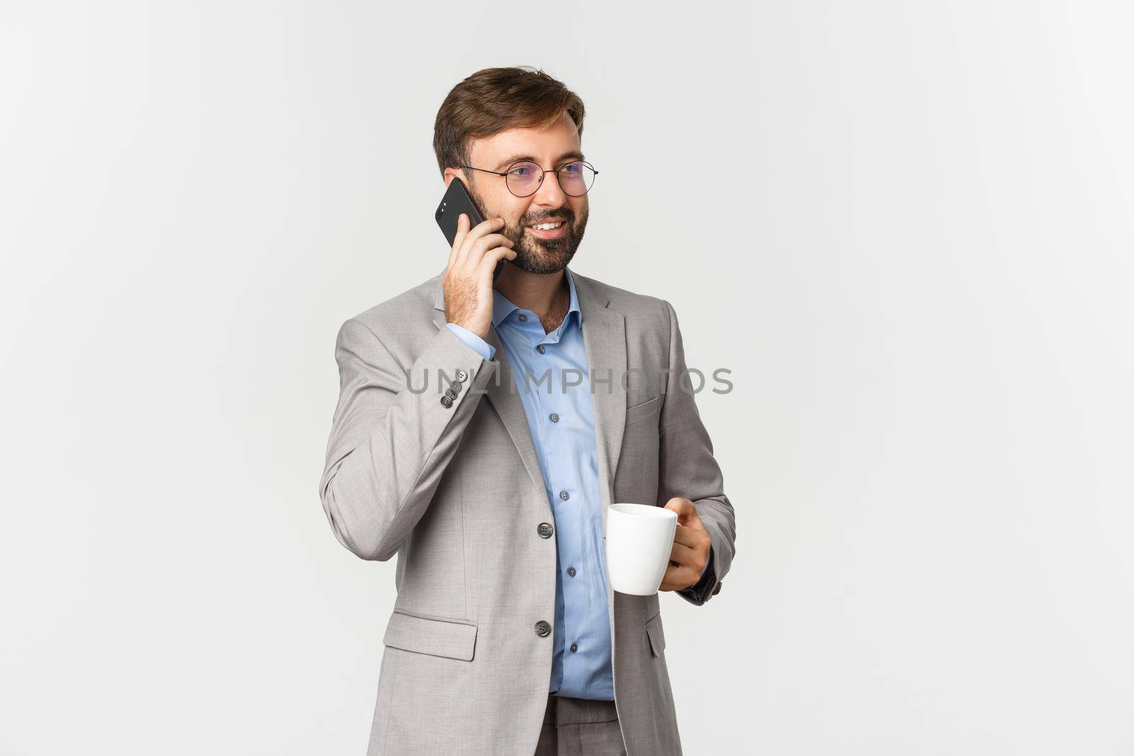 Image of successful businessman in grey suit and glasses, drinking coffee and talking on phone with pleased smile, standing over white background by Benzoix