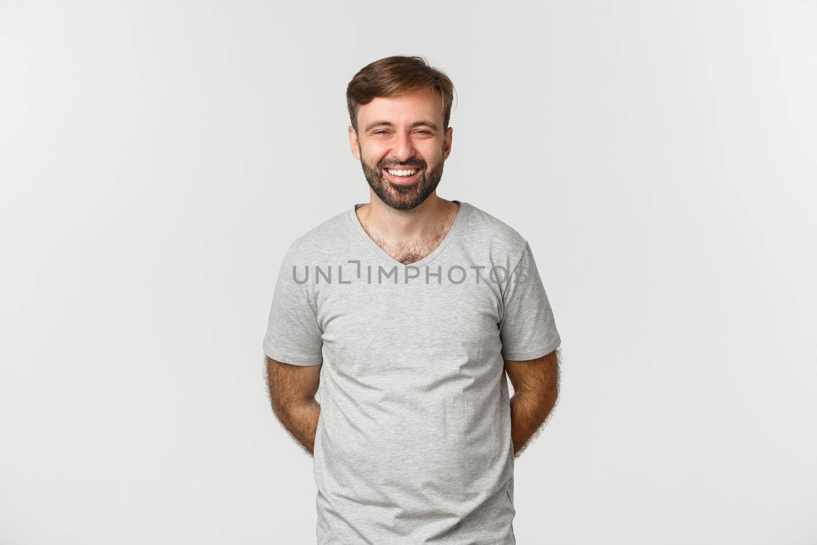 Image of happy bearded man in gray t-shirt, smiling and laughing, standing humble with hands behind.