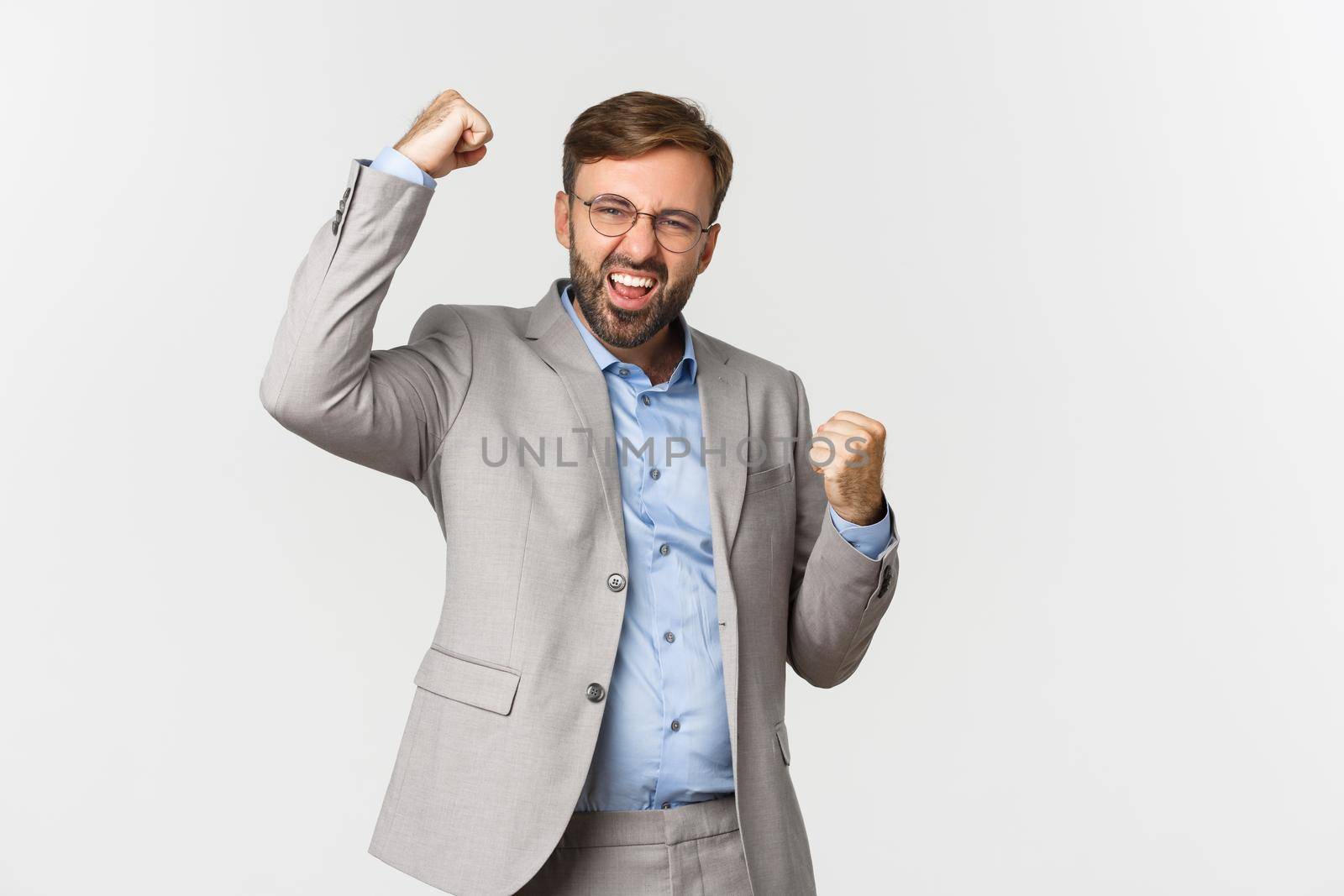 Portrait of successful bearded businessman in grey suit, achieve goal and triumphing, making fist pump gesture and saying yes with confident look, standing over white background.