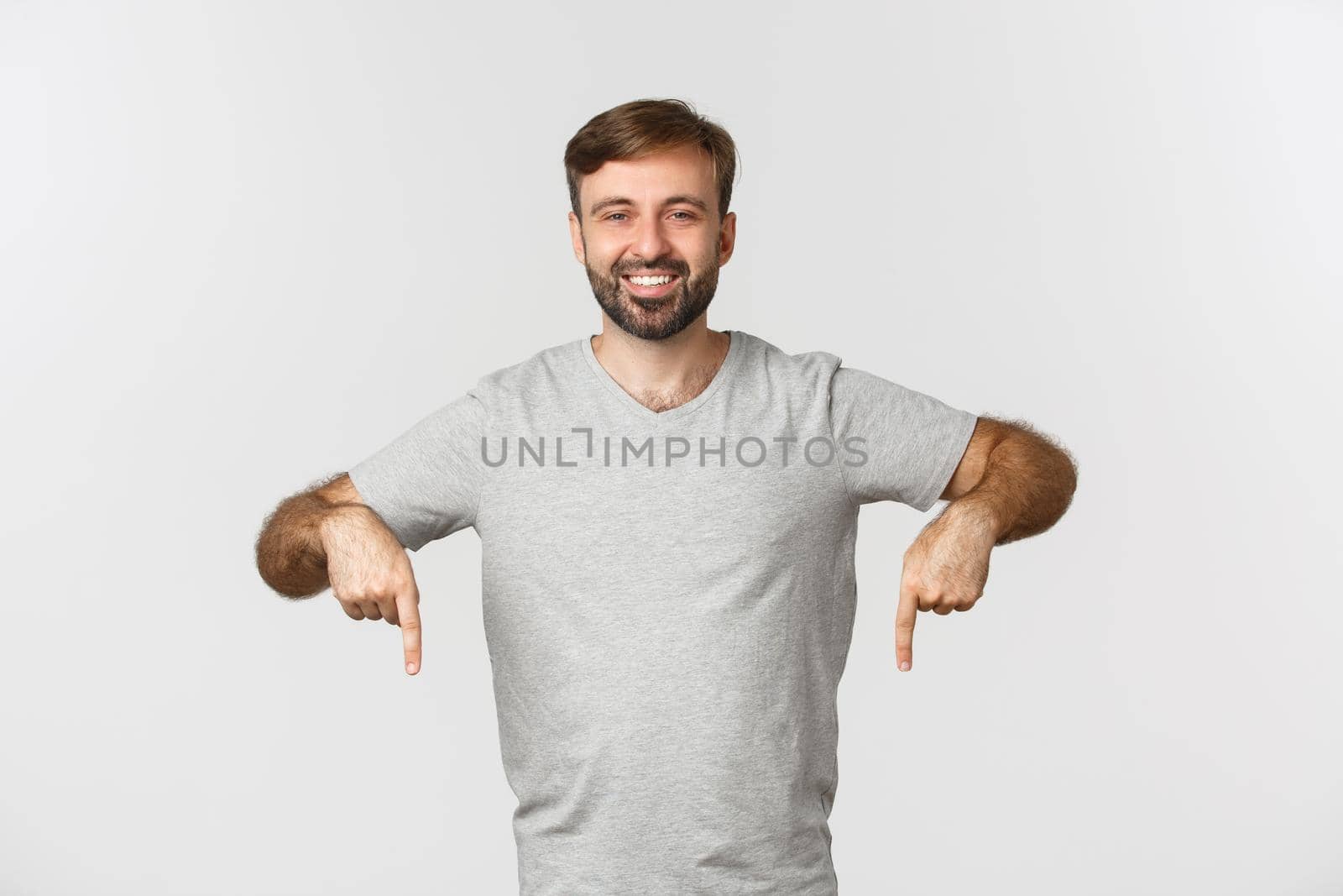 Portrait of attractive bearded man in gray t-shirt, showing logo, pointing fingers down, standing over white background.