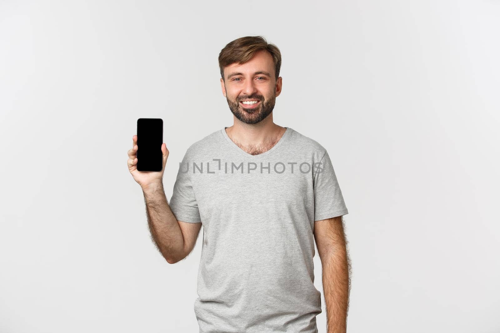 Portrait of smiling handsome man in gray t-shirt, showing mobile phone screen, recommending app or shopping site, standing over white background.