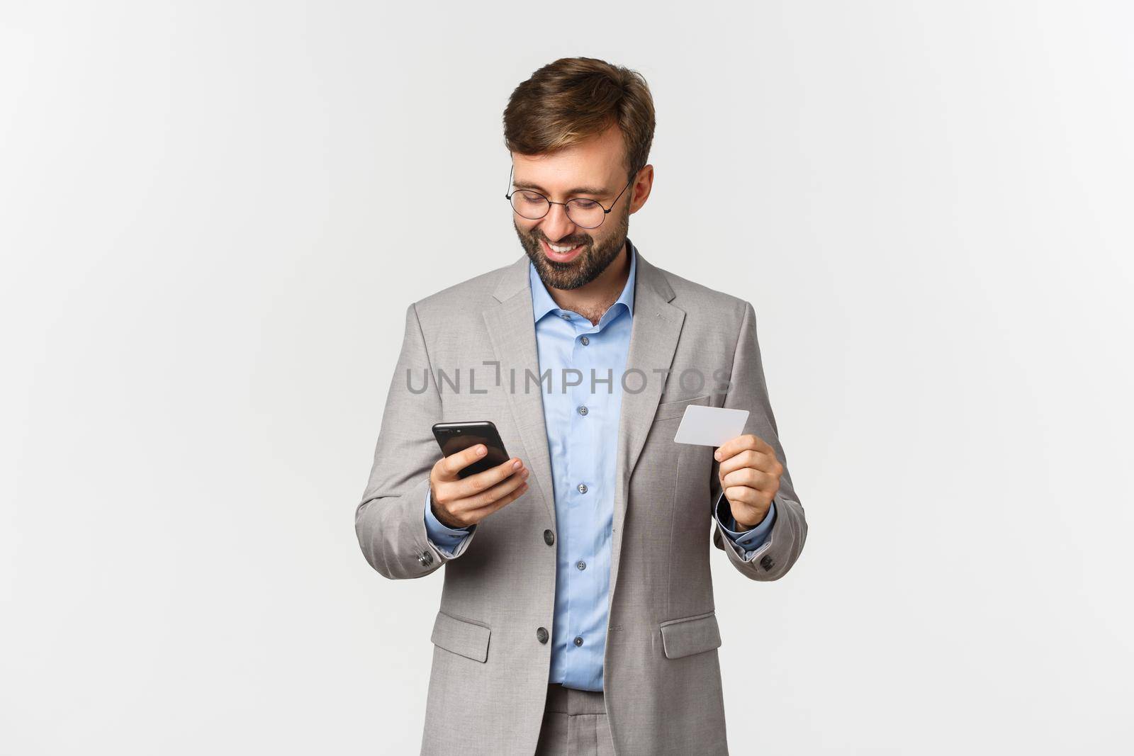 Handsome bearded businessman in grey suit and glasses, shopping online, holding credit card and mobile phone, standing over white background.