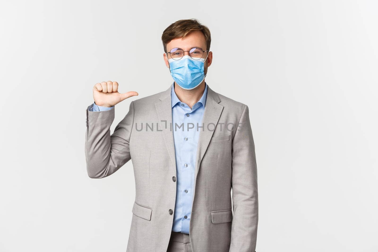 Concept of work, covid-19 and social distancing. Happy businessman in glasses, grey suit and medical mask pointing at himself, showing example to employees, standing over white background by Benzoix