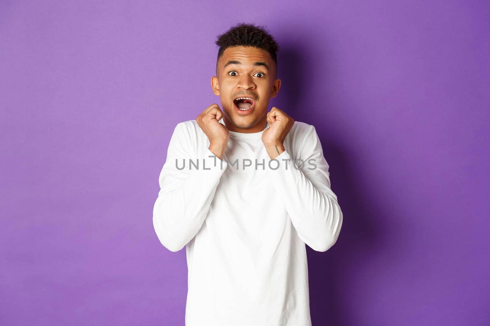 Portrait of astonished african-american guy in white casual shirt, open mouth fascinated and looking at cool promo, standing over purple background.