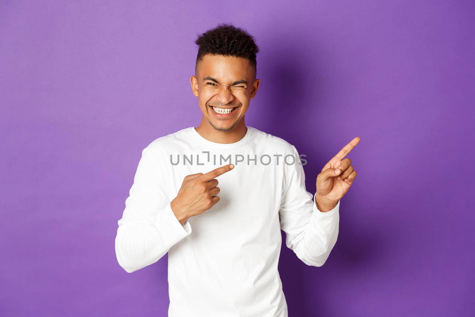 Image of handsome and carefree african-american man in white sweatshirt, pointing fingers right and laughing at something funny, standing over purple background.