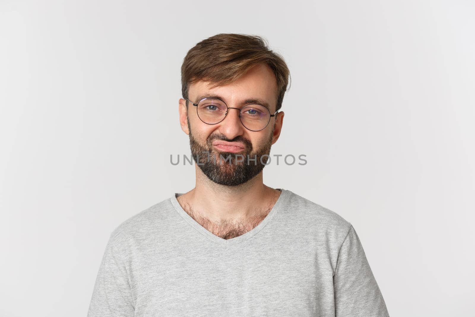 Close-up of skeptical bearded man in glasses and gray t-shirt, grimacing reluctant, standing over white background.