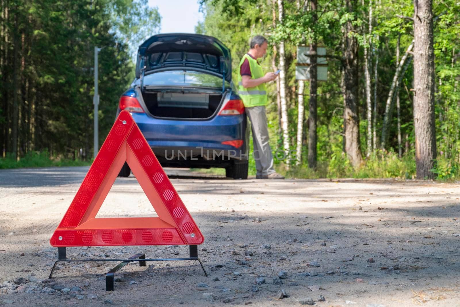 Close-up of a portable reflective red warning triangular sign on the side of a rural road near a car. The driver on the phone calling for technical assistance. Selective focus.