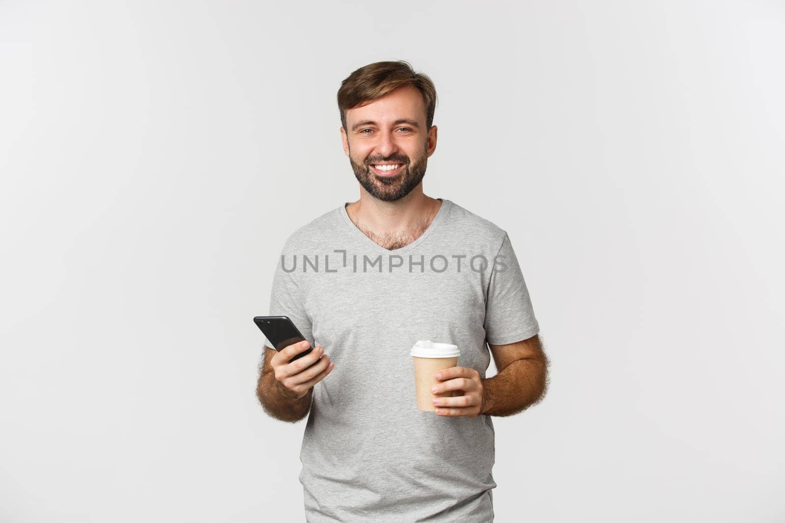 Image of modern caucasian man with beard, looking happy while drinking coffee and casually using mobile phone, standing over white background.