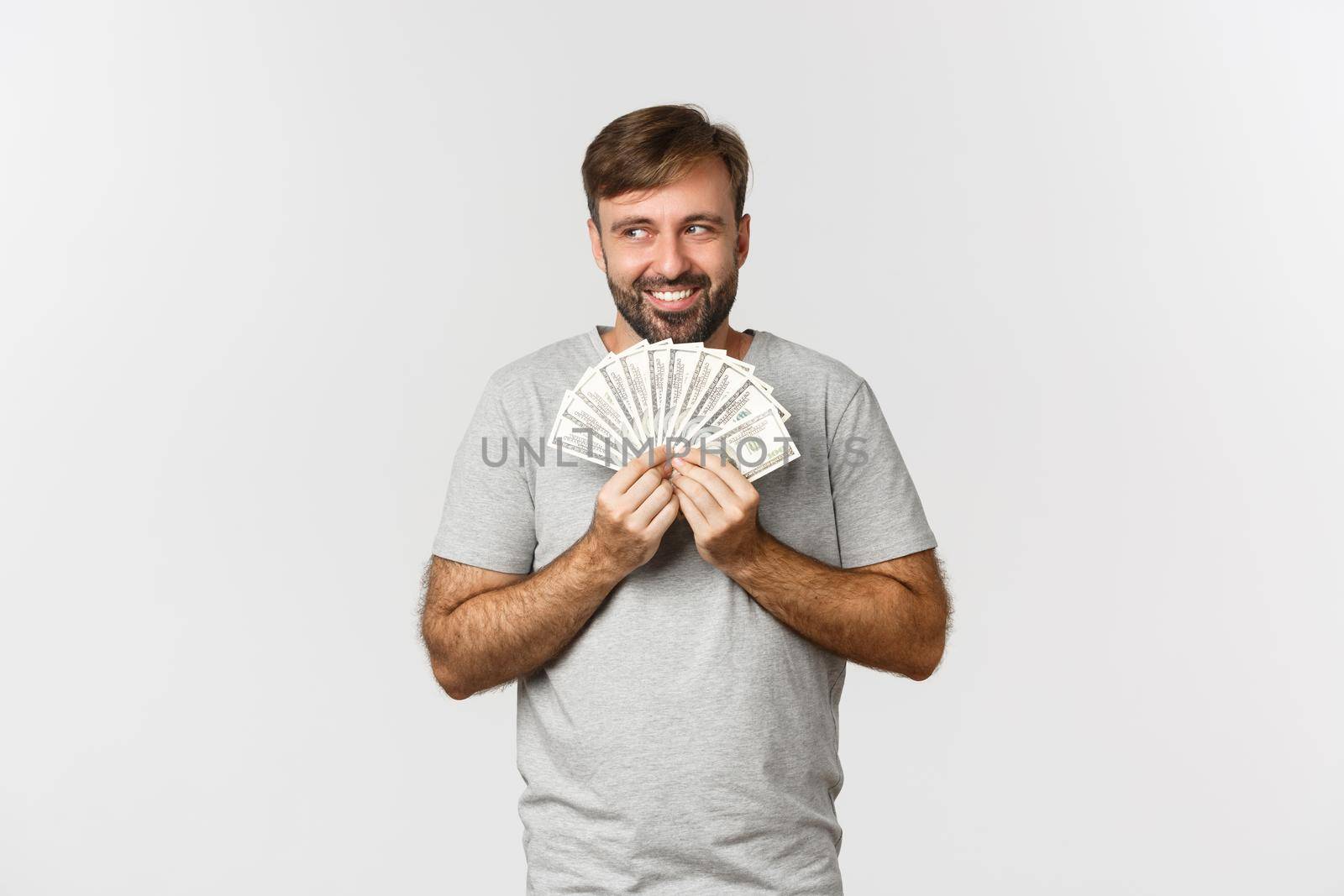 Greedy smiling greedy man with beard, thinking about shopping, holding money and looking at upper left corner, standing over white background.