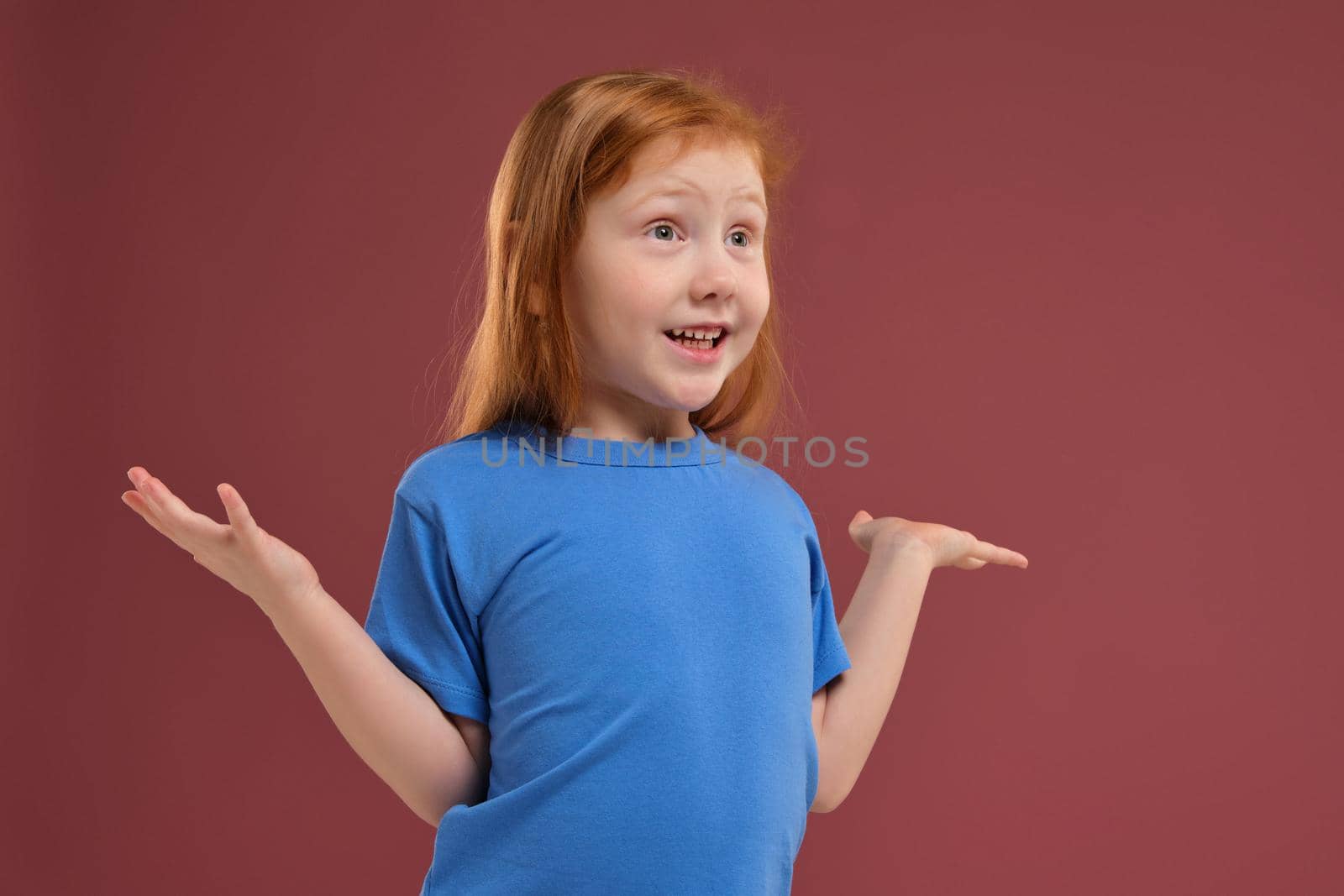 Portrait of cute redhead emotional little girl in blue t-shirt on red background. Beautiful studio portrait of female little caucasian person spreading her hands, she's so happy