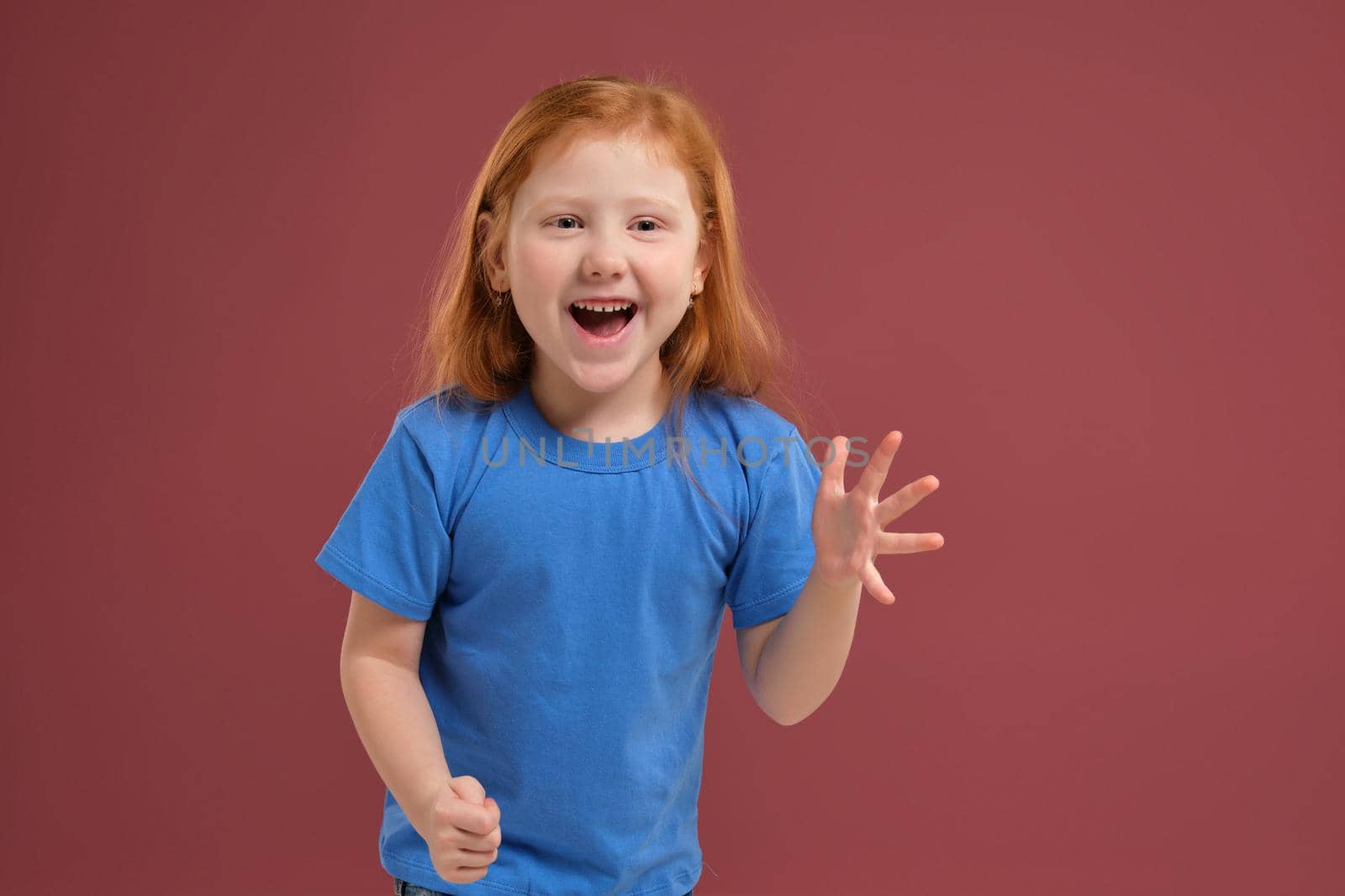 Portrait of cute redhead emotional little girl in blue t-shirt on red background. Beautiful studio portrait of female little caucasian person spreading her hands, she's so happy