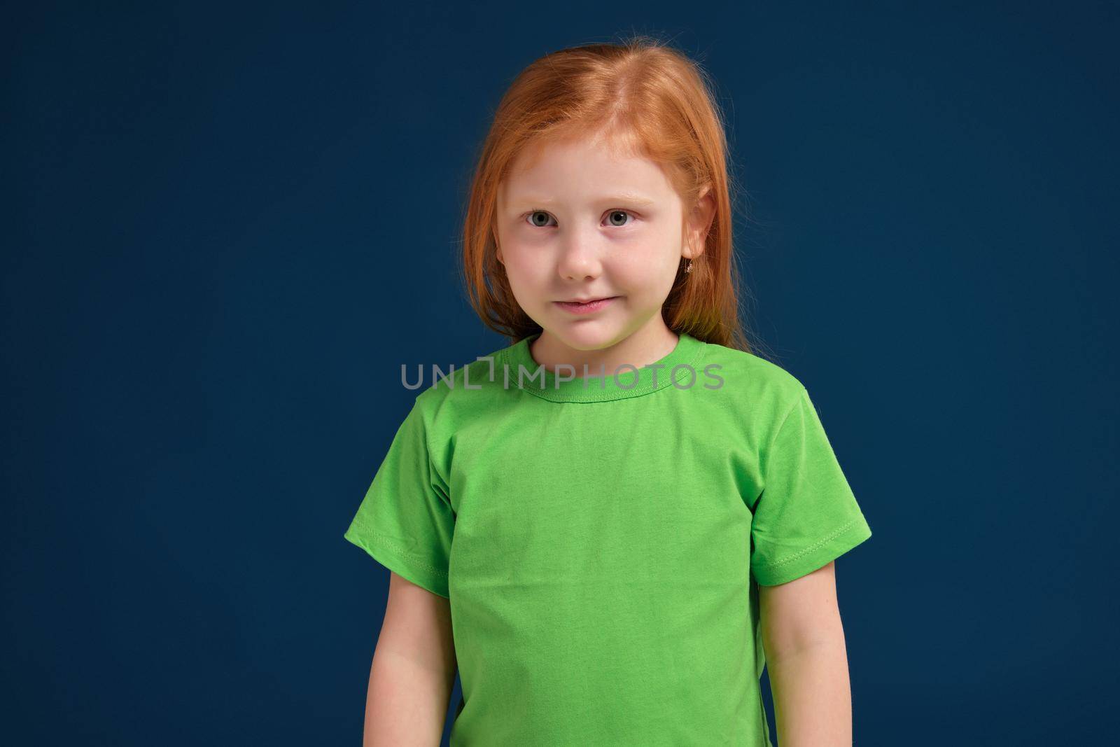 Portrait of cute redhead emotional little girl in green t-shirt on blue background. Beautiful studio portrait of female little caucasian person. She looks so happy and shy a little bit