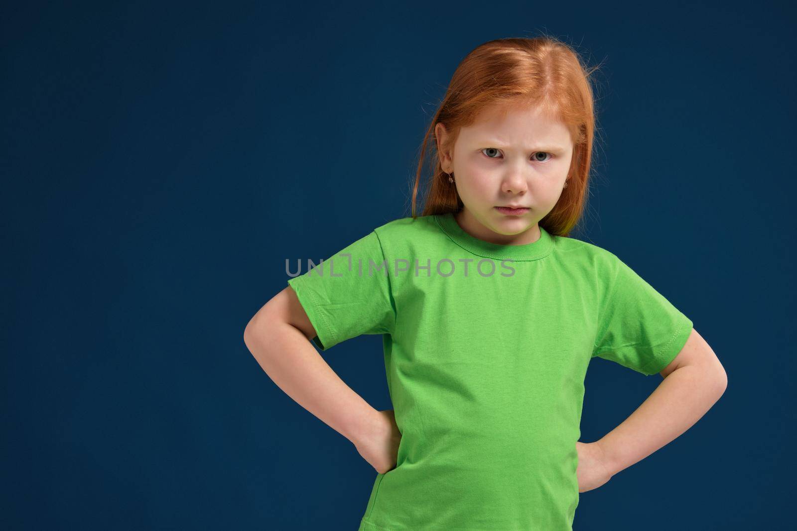 Portrait of angry offended displeased redhead little girl, holding hands on hips, looking from under forehead