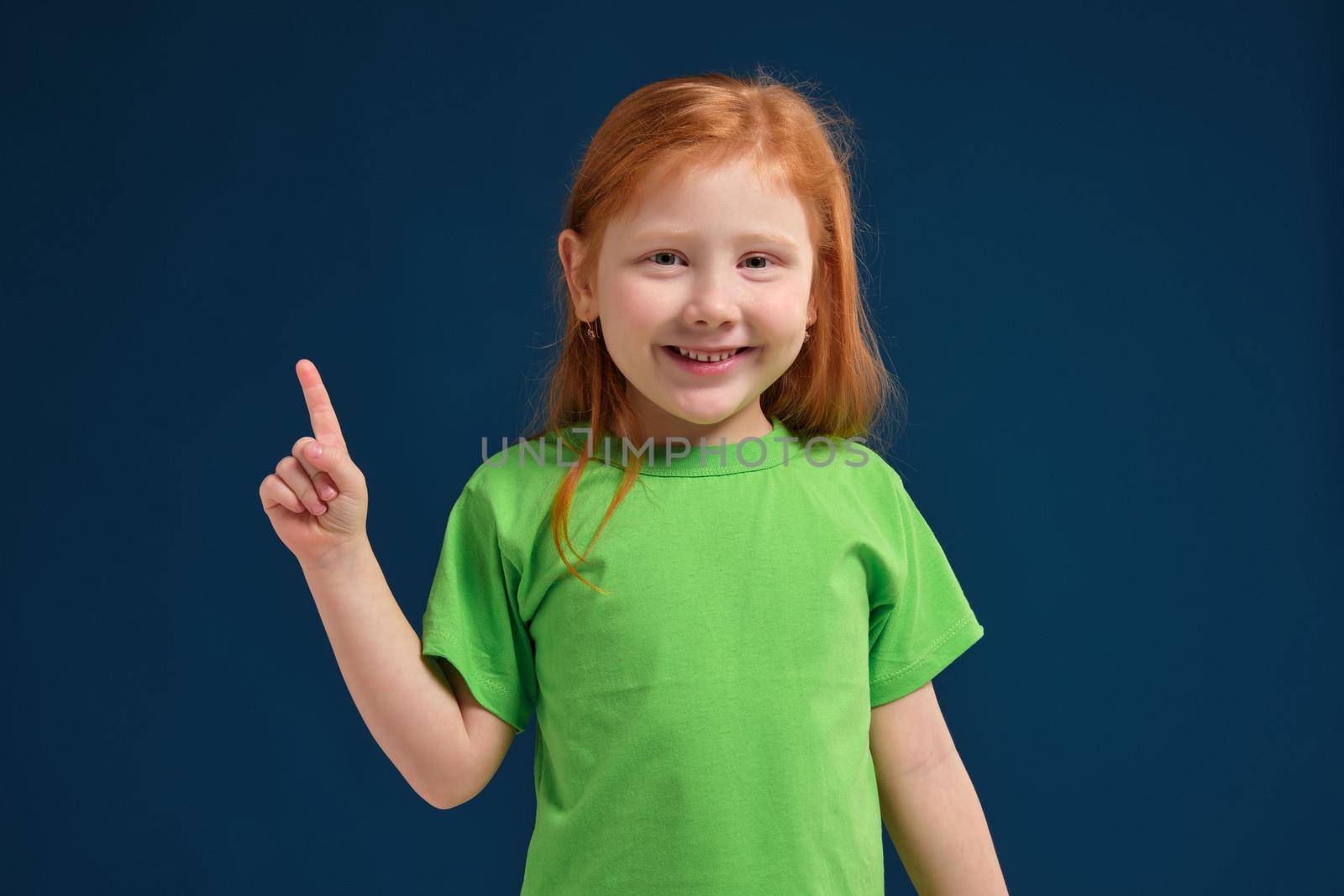 close up view of adorable happy redhead little girl in green t-shirt on blue background pointing up with her finger, smiling
