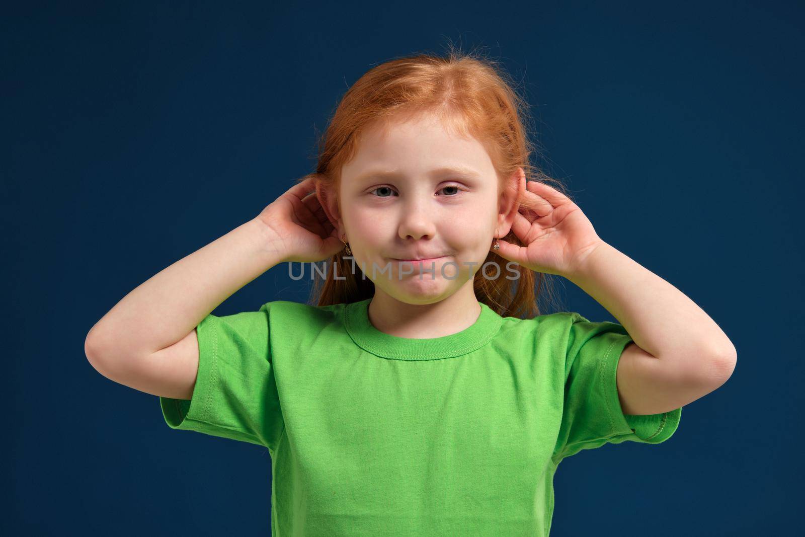 Portrait of cute redhead little girl trying to hear someone putting her hand on the ear, blue background, green t-shirt