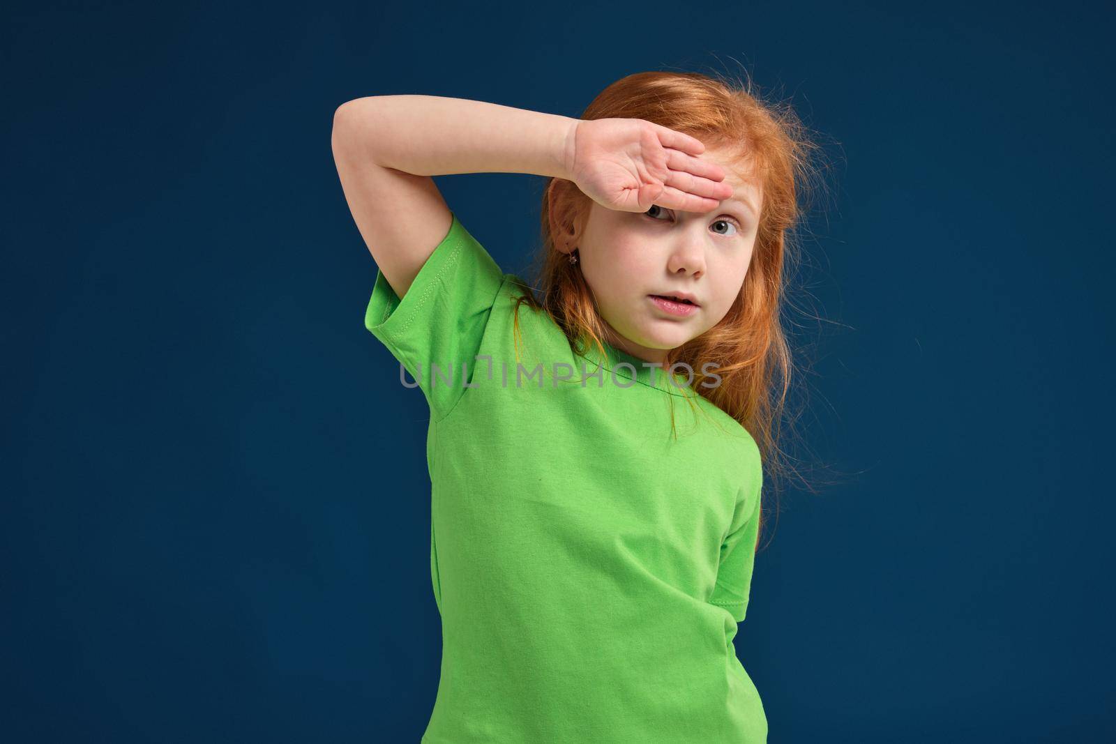 Funny photo of little redhead girl covering her eyes up from bright light or sun, blue background, green t-shirt