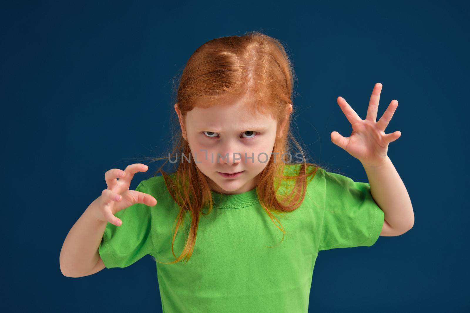 Little redhead cute girl trying to frighten someone with stretched hands, green t-shirt, blue background