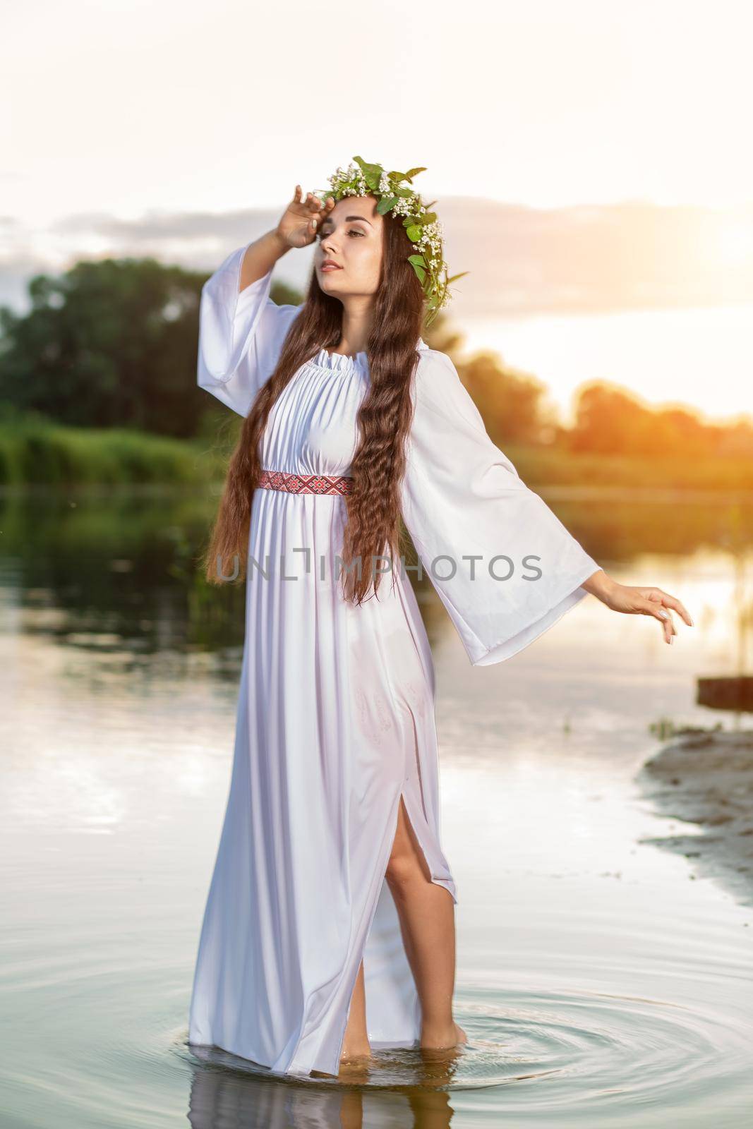 Beautiful black haired girl in white vintage dress and wreath of flowers standing in water of lake. Sun flare. by nazarovsergey