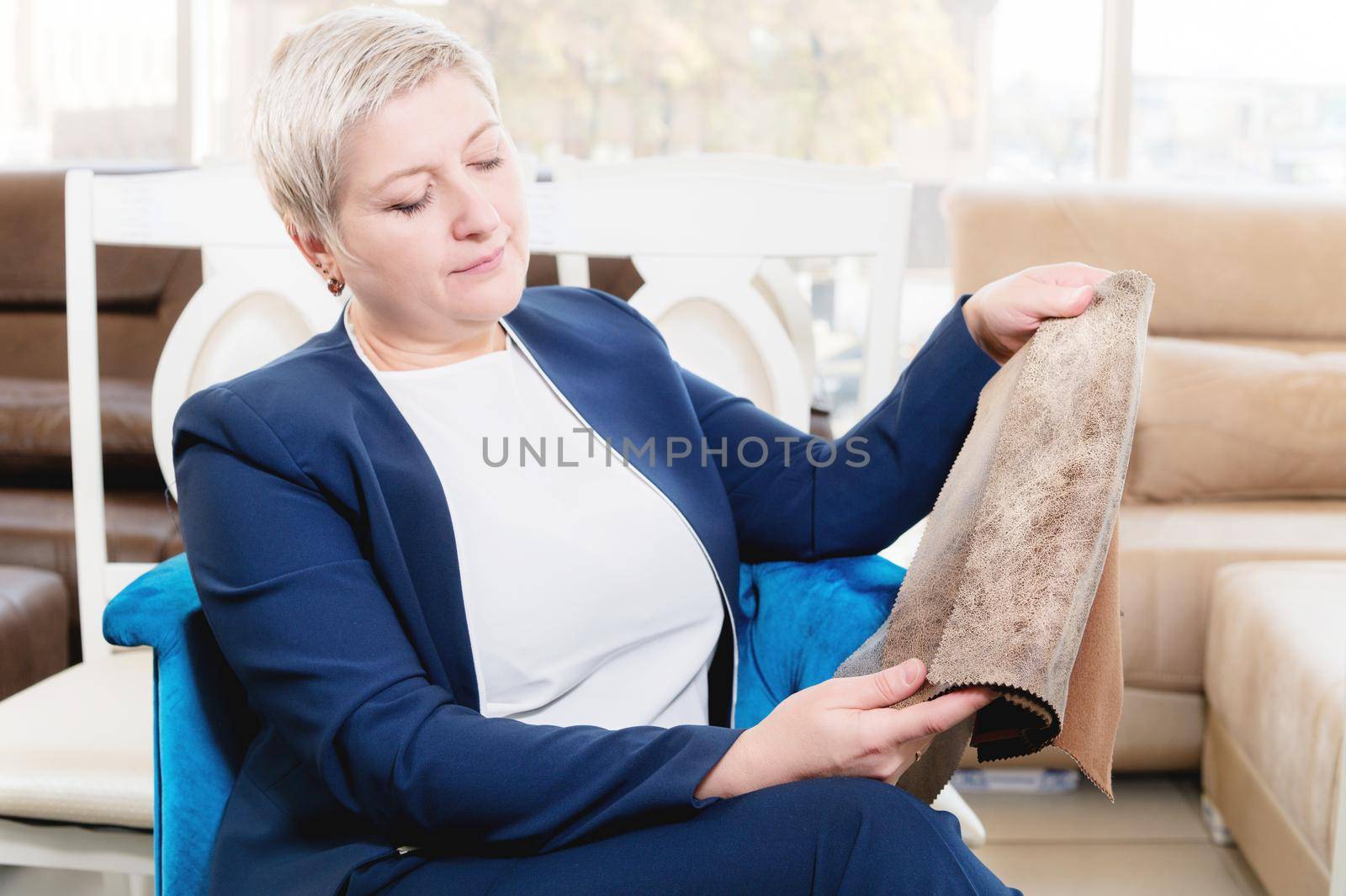 Portrait of an attractive mature short-haired woman in a pink sweater sits in a leather armchair in a furniture store and selects the color and upholstery material of the furniture. by yanik88