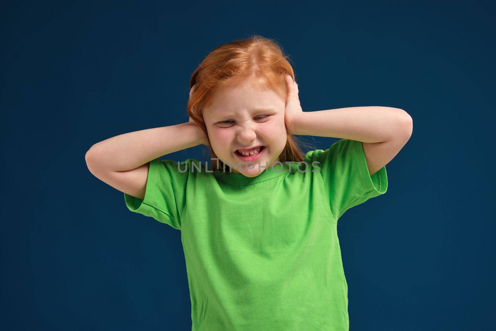 Cute young little redhead girl in green t-shirt covering her ears with her hands, narrowed eyes, blue background