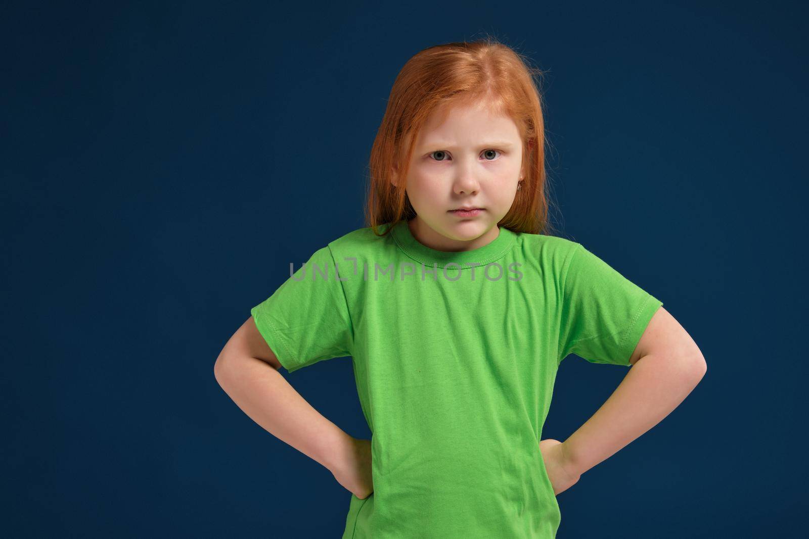 Portrait of angry offended displeased redhead little girl, holding hands on hips, looking from under forehead