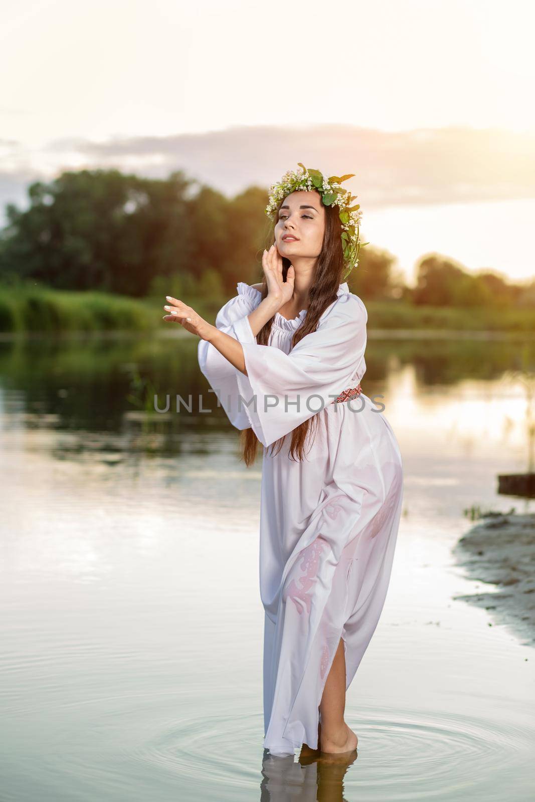 Beautiful black haired girl in white vintage dress and wreath of flowers standing in water of lake. Fairytale story. Warm art work. Sun flare