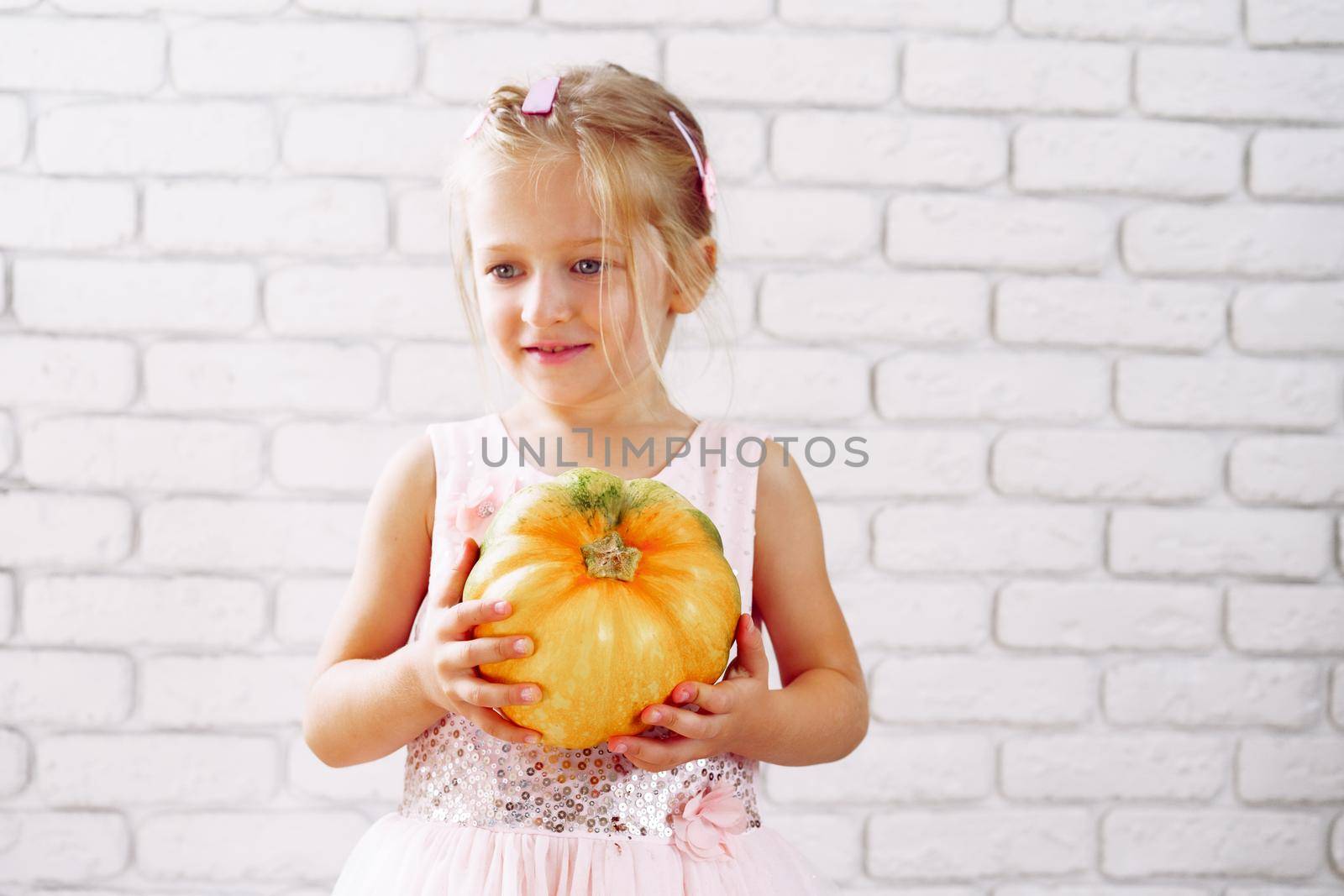 Cute little girl in pink dress holding pumpkin in her hands against white brick wall