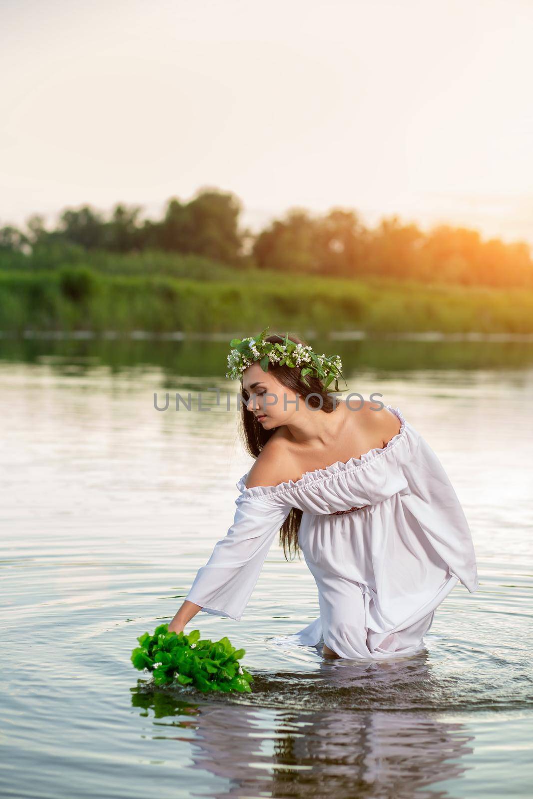 Beautiful black haired girl in white vintage dress and wreath of flowers standing in water of lake. Fairytale story. Warm art work. Sun flare