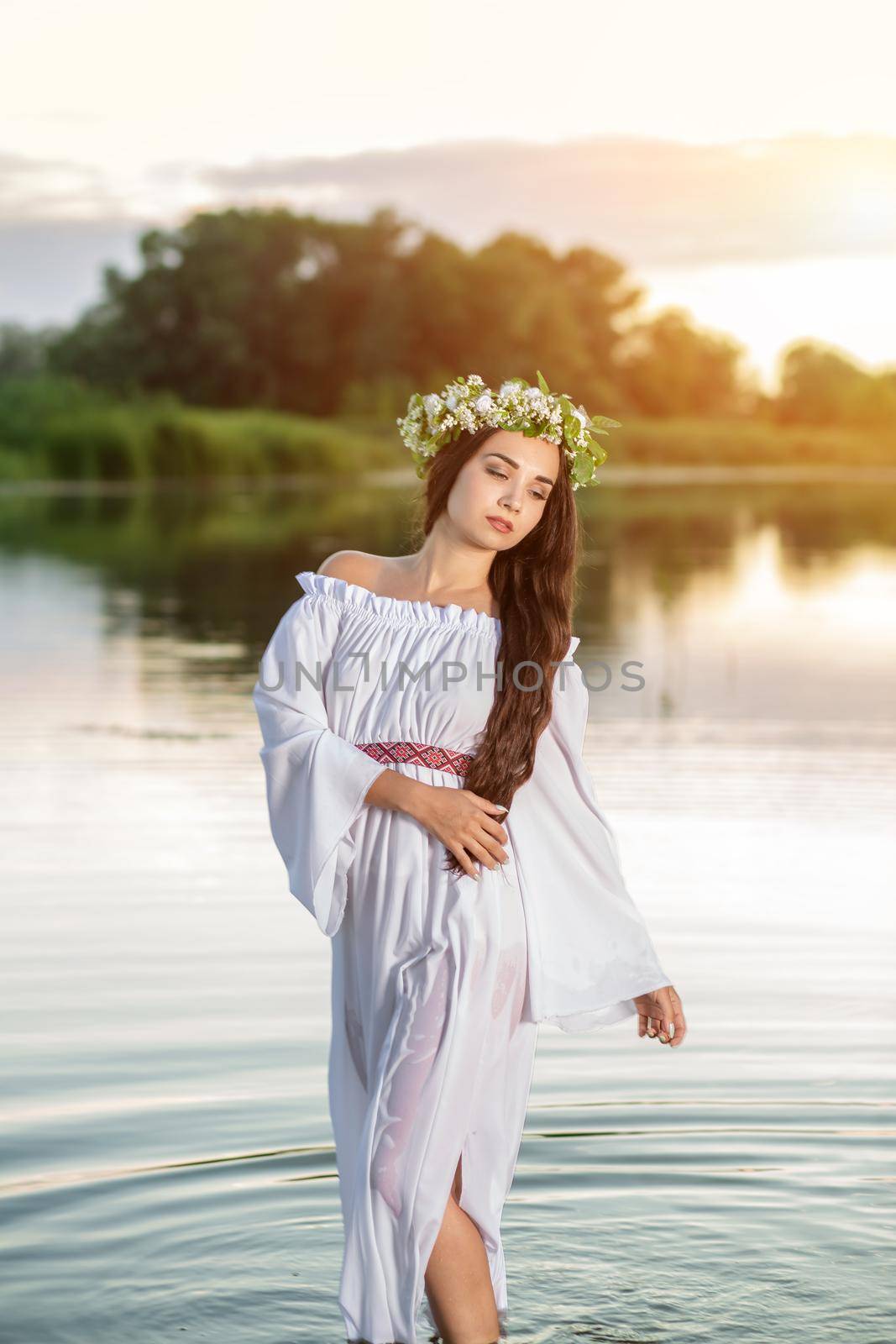 Beautiful black haired girl in white vintage dress and wreath of flowers standing in water of lake. Fairytale story. Warm art work. Sun flare