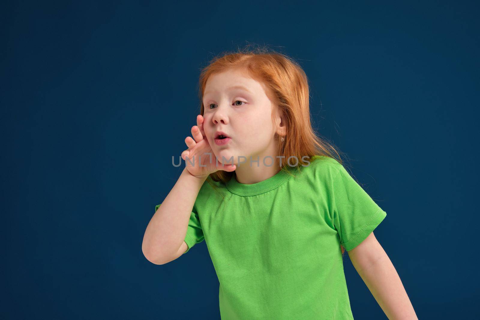 Portrait of a small pretty redhead girl standing and calling someone holding her hand near her mouth wearing a green t-shirt ,blue background