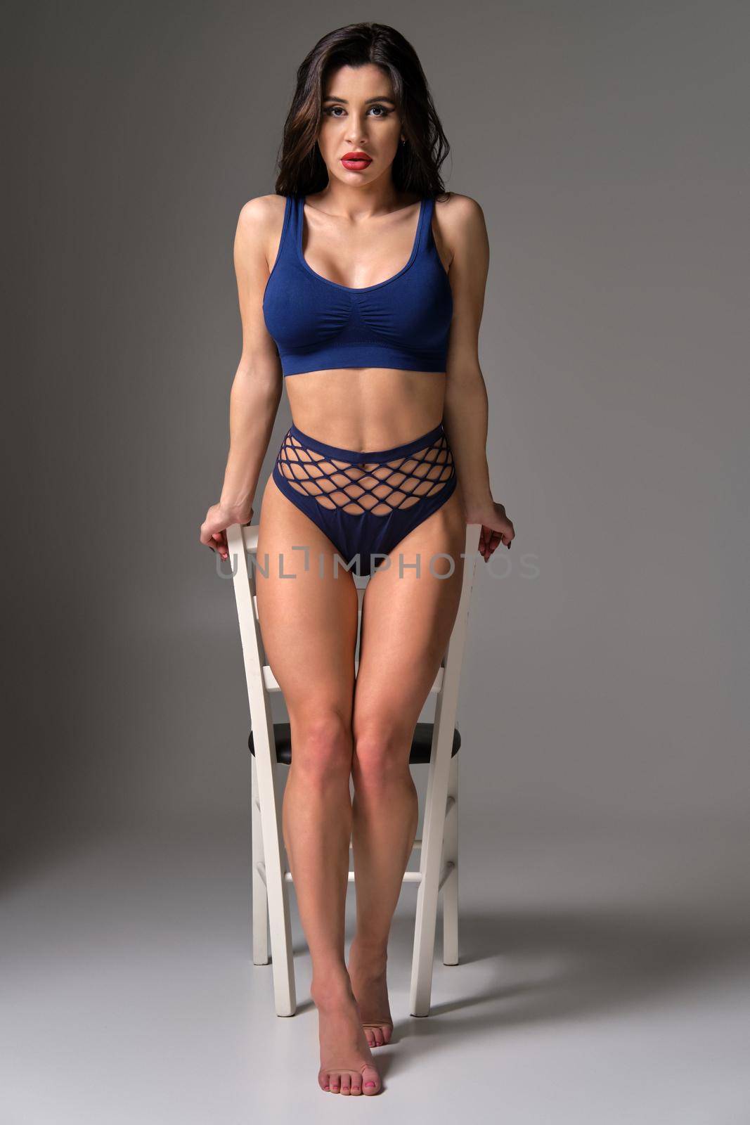 Full length studio shot of a glamour brunette lady in a sexy lingerie posing against a white chair on a gray background. Seductive model with a bright make-up and in a blue underwear. Fashionable lifestyle, sensuality and beauty concept.