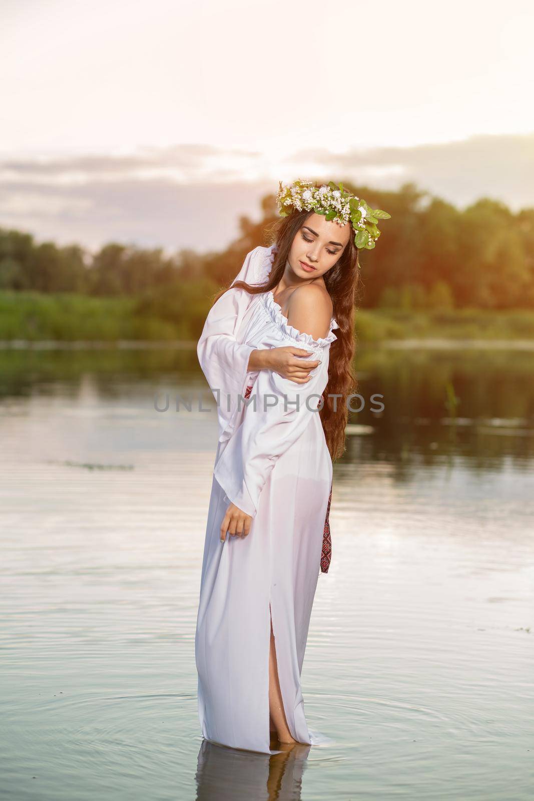 Beautiful black haired girl in white vintage dress and wreath of flowers standing in water of lake. Sun flare. by nazarovsergey