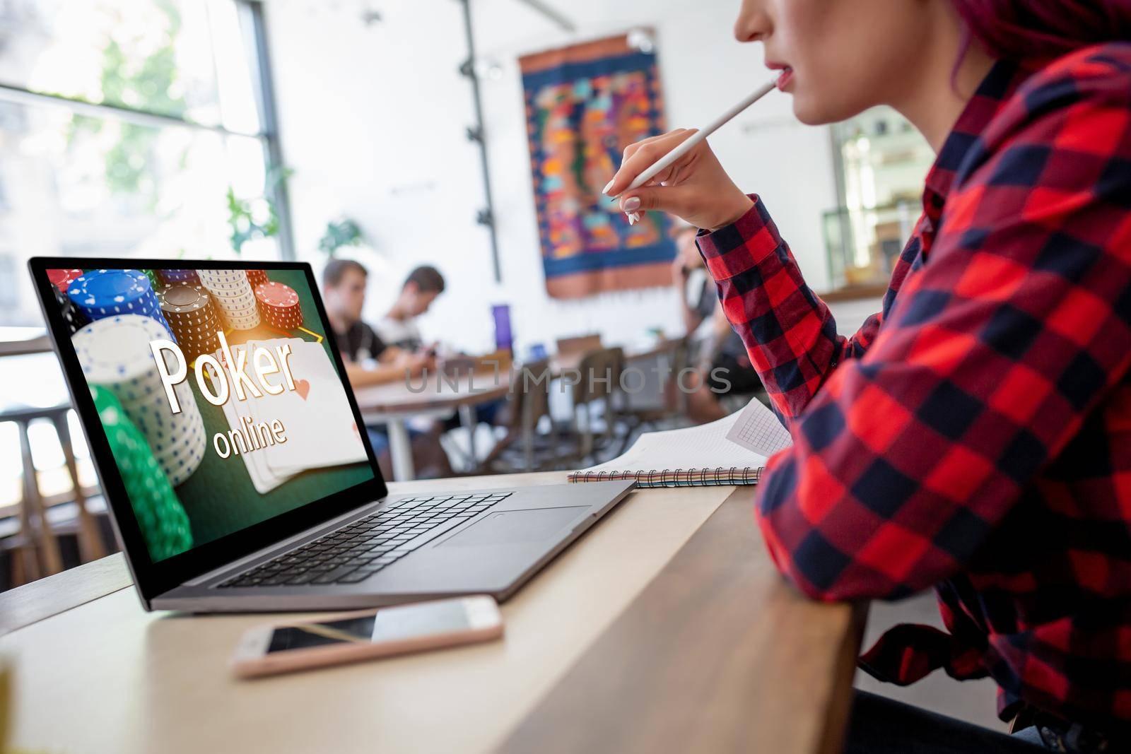 Side view of a young pink hair woman keyboarding on laptop computer with poker online on a screen while sitting in cafe. Play online concept. Screen graphics are made up.