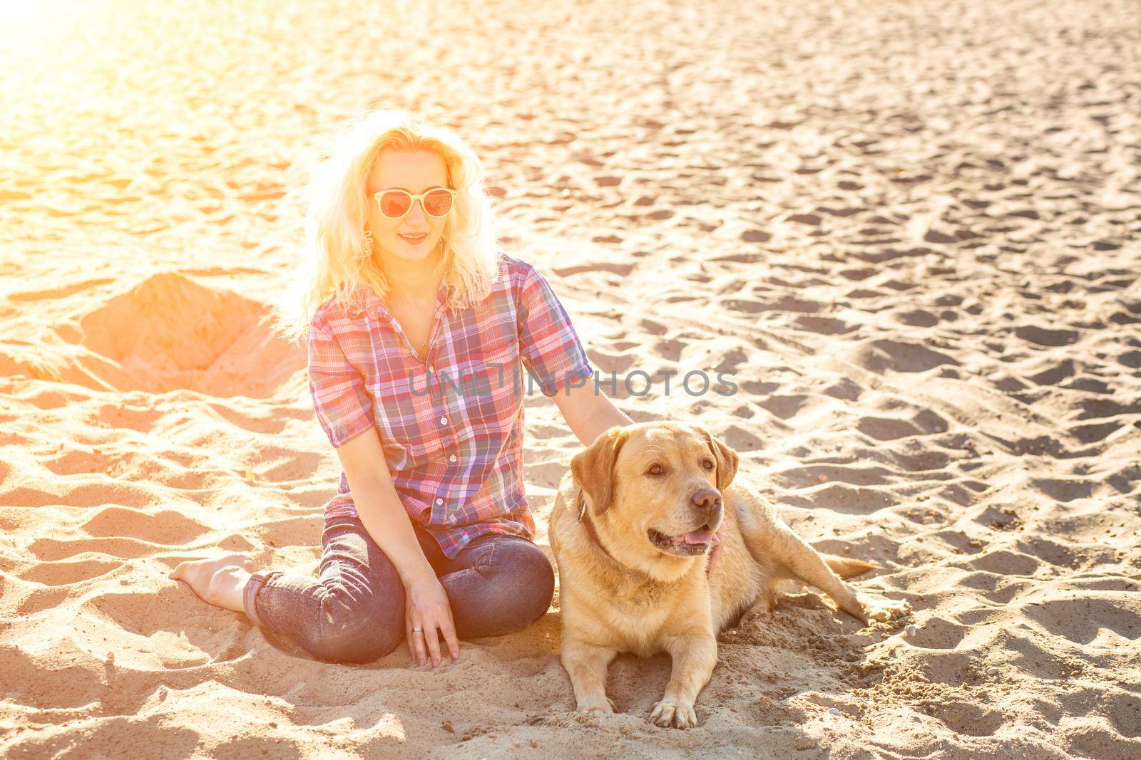 Portrait of young beautiful woman in sunglasses sitting on sand beach with golden retriever dog. Girl with dog by sea. Happiness and friendship. Pet and woman. Woman playing with dog on sea shore. Sun flare
