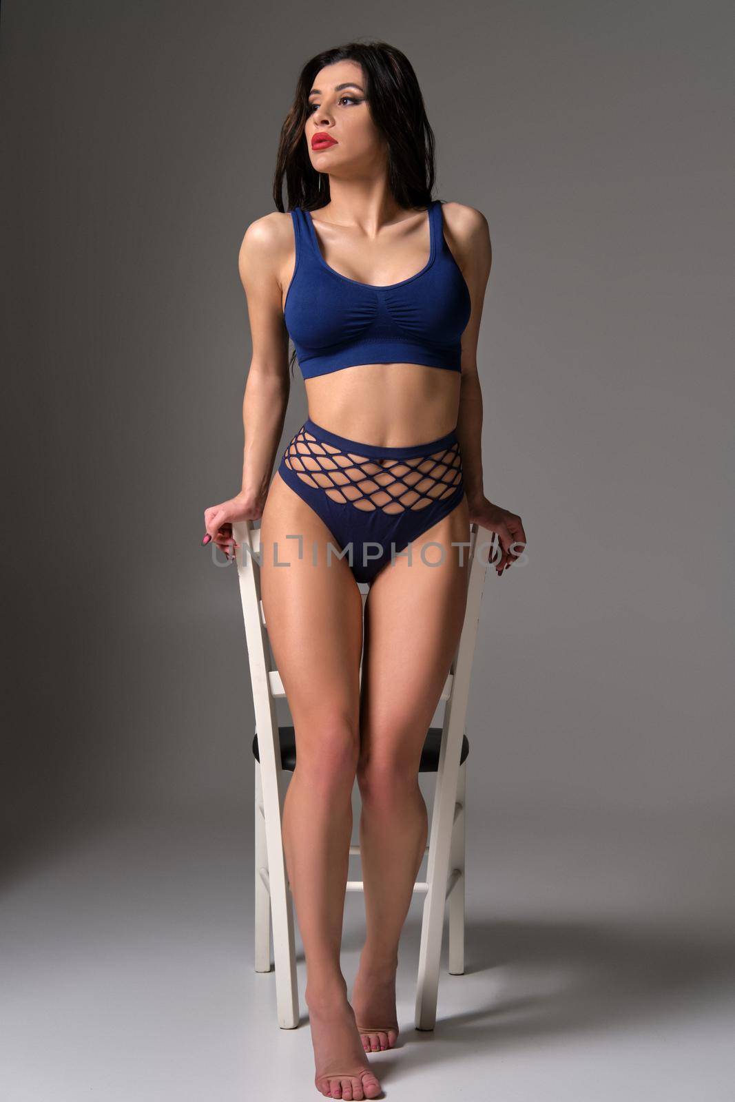 Full length studio shot of a glamour brunette maiden in a sexy lingerie looking away while posing against a white chair on a gray background. Seductive model with a bright make-up and in a blue underwear. Fashionable lifestyle, sensuality and beauty concept.