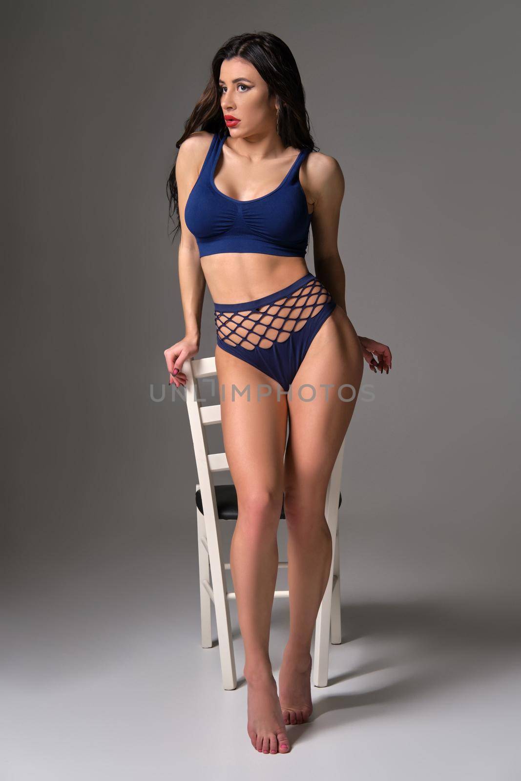 Full length studio shot of an attractive brunette woman in a sexy lingerie looking away while posing against a white chair on a gray background. Young model with a bright make-up and in a blue underwear. Fashionable lifestyle, sensuality and beauty concept.