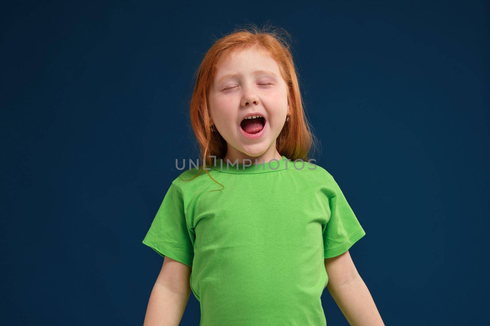 close up photo of little redhead emotional girl in green t-shirt posing before camera. She is tired and wants to sleep, yawning and stretching