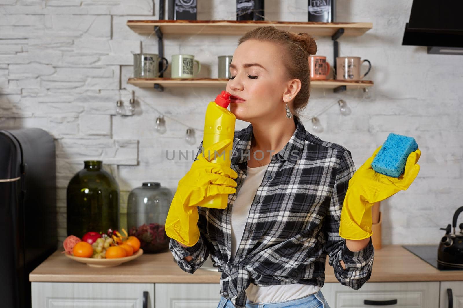 Housekeeper concept. Professional young woman wearing rubber protective yellow gloves holding bottle cleaners in the kitchen. Housewife hold bottle spray work clean at home.