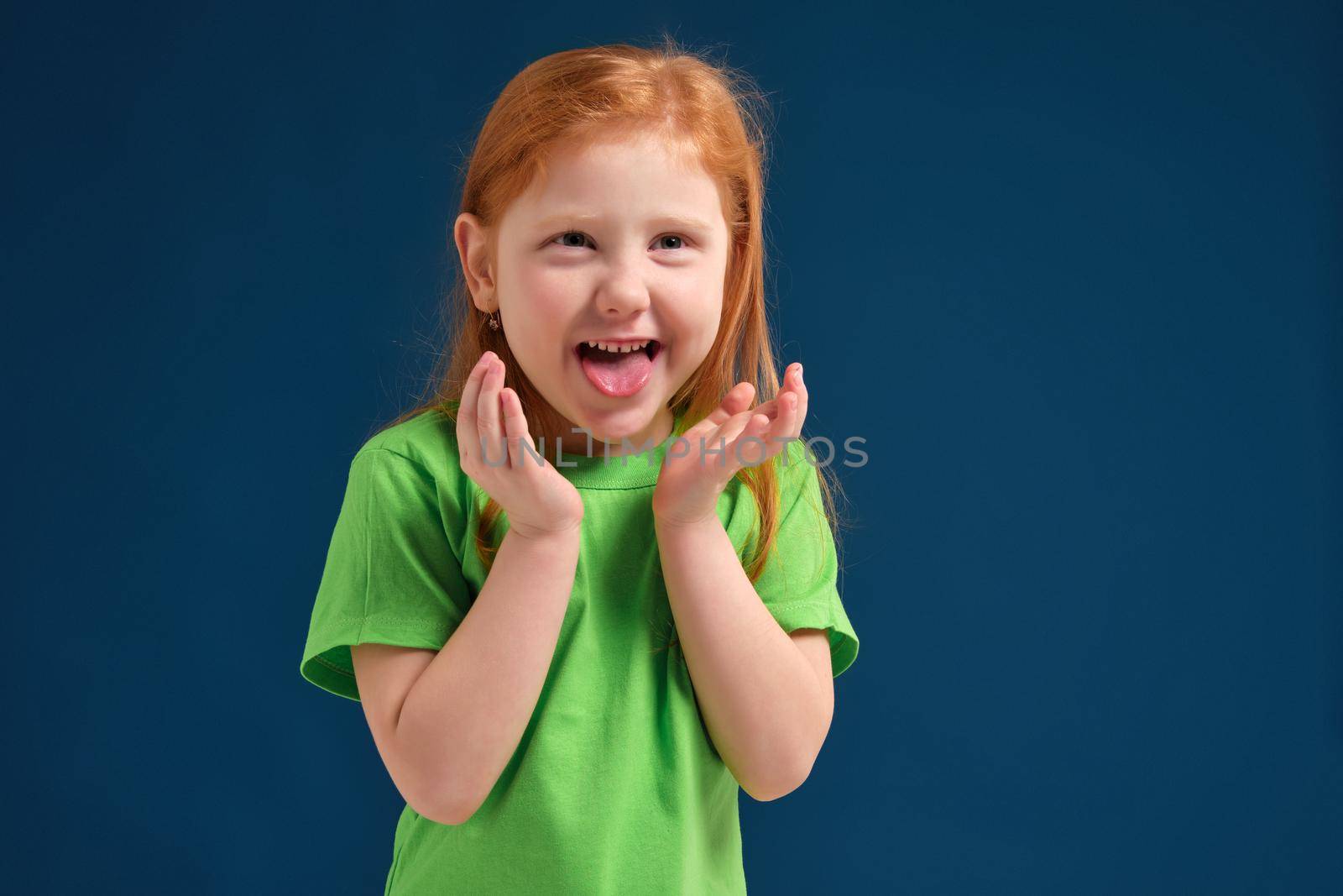 Portrait of cute redhead emotional little girl in green t-shirt on blue background. Beautiful studio portrait of female little caucasian personh holding her hands under her chin, she's looks happy