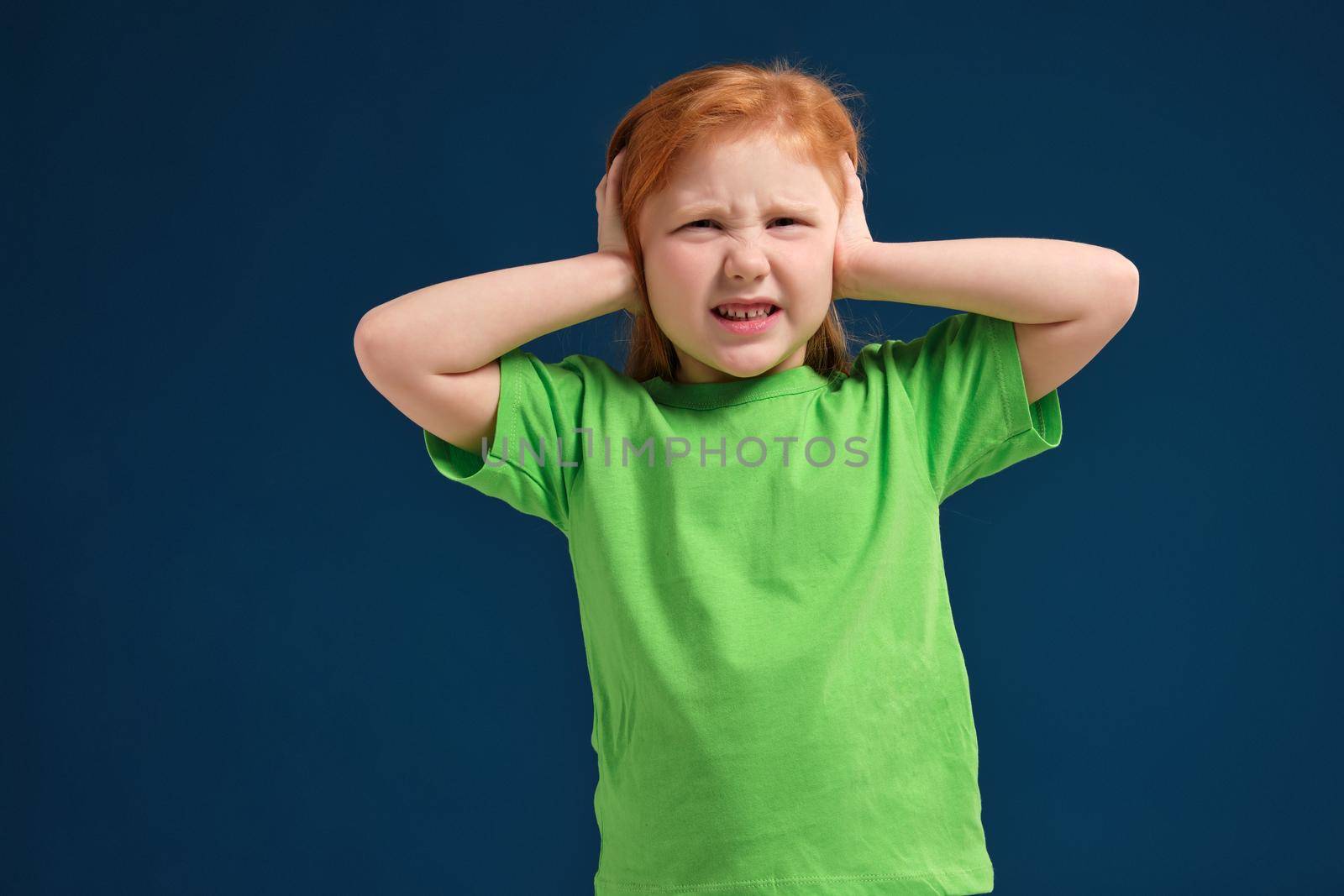 Cute young little redhead girl in green t-shirt covering her ears with her hands, narrowed eyes, blue background
