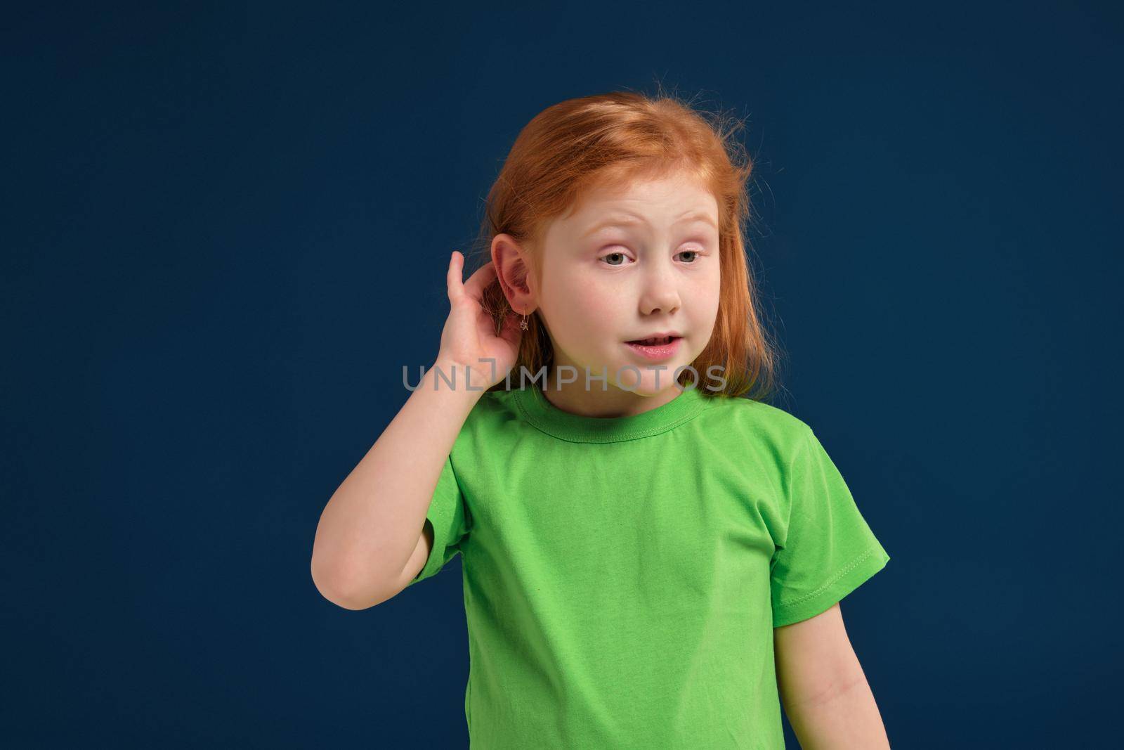 Portrait of cute redhead little girl trying to hear someone putting her hand on the ear, blue background, green t-shirt