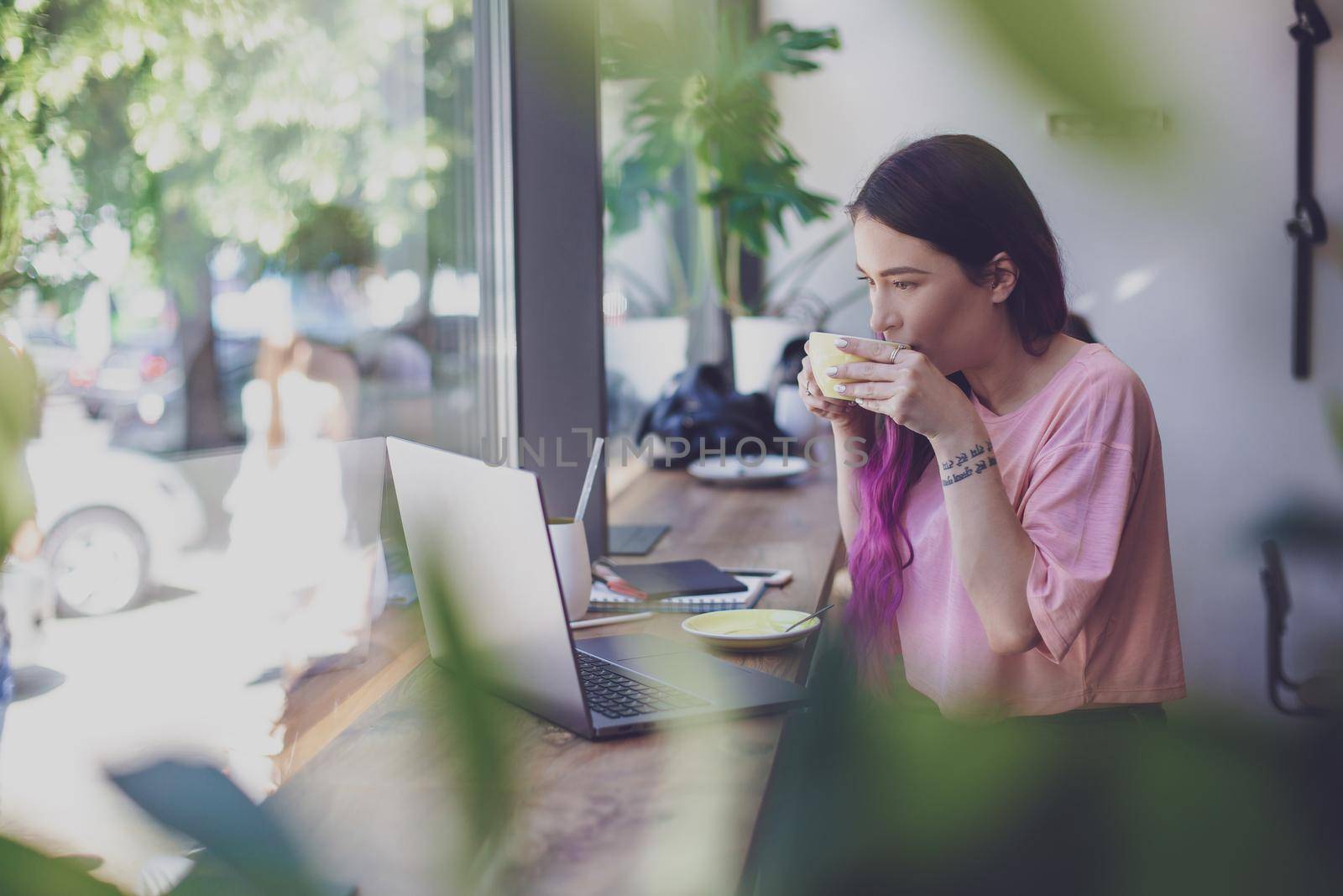 Side view of young businesswoman sitting at table in coffee shop. On table cup of coffee and laptop. In background white wall and window. Girl shopping online.