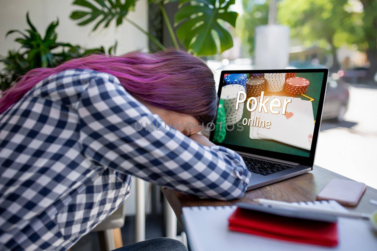 Side view of a young pink hair woman keyboarding on laptop computer with poker online on a screen while sitting in cafe. Play online concept. Screen graphics are made up. Emotions. Loss