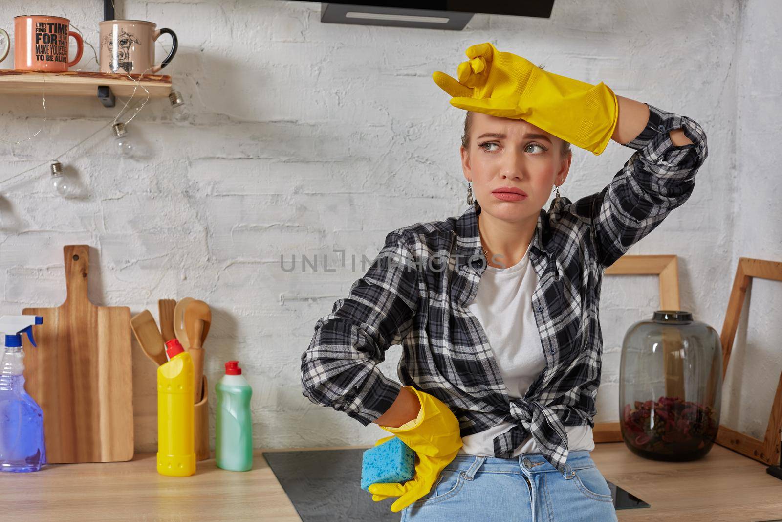Blonde woman in protective gloves with rag cleaning electric stove at home kitchen. Girl washing black shiny surface of kitchen top. Concept of housework.