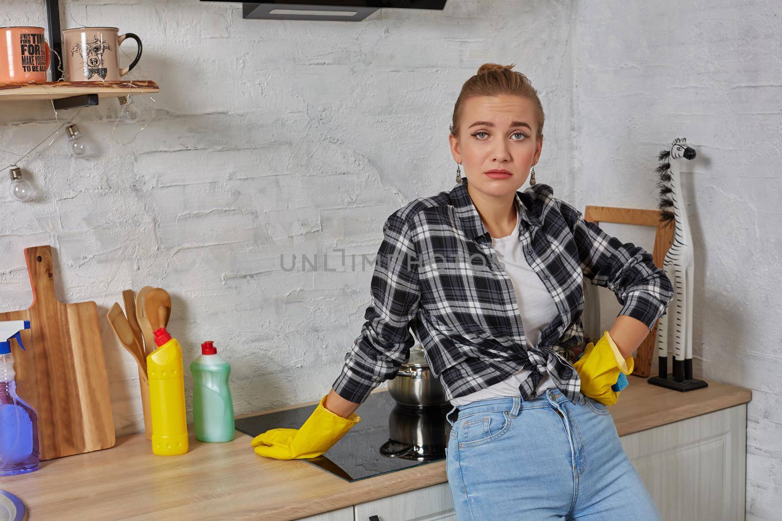 Blonde woman in protective gloves with rag cleaning electric stove at home kitchen. Girl washing black shiny surface of kitchen top. Concept of housework.