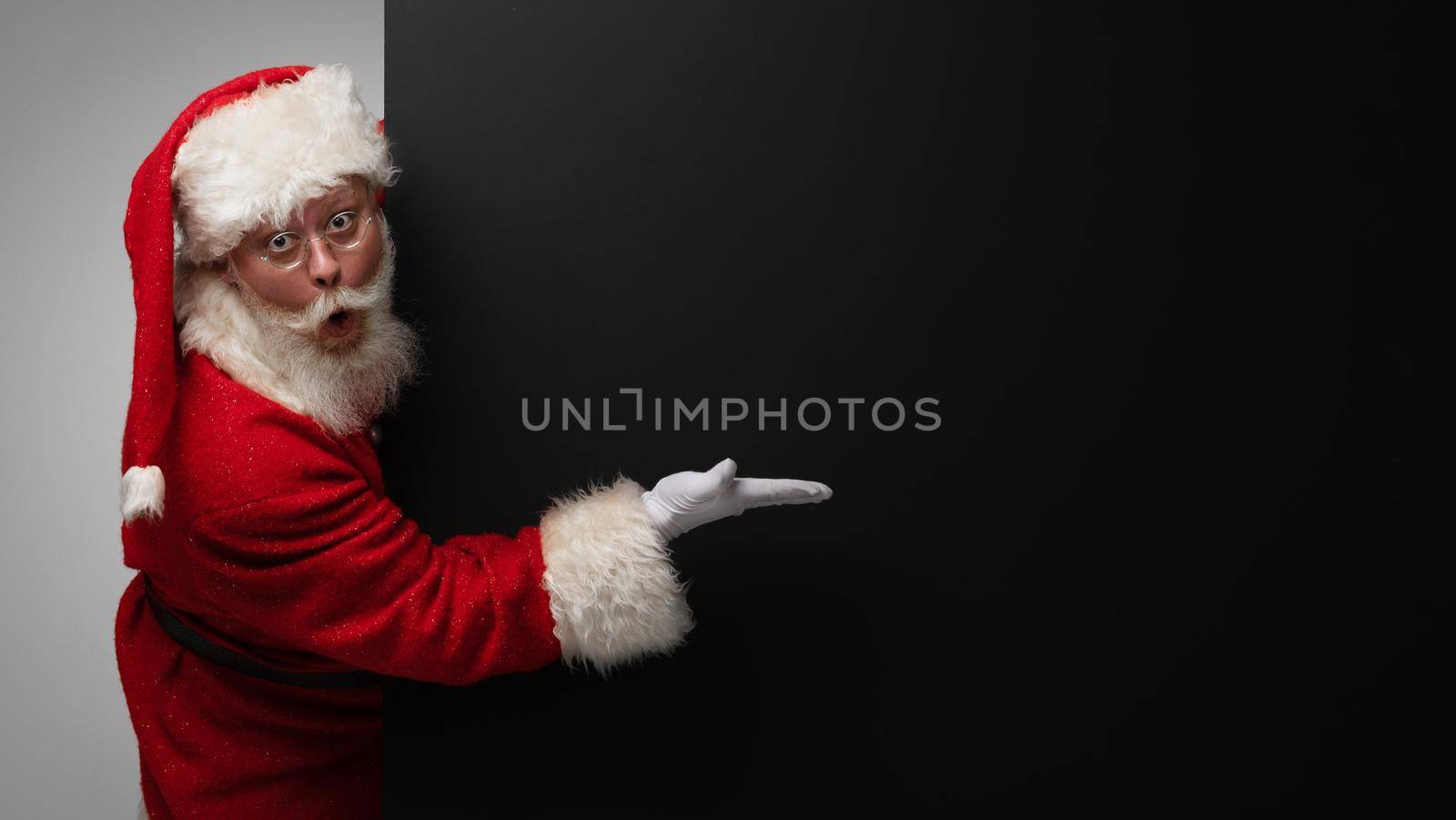 Santa Claus pointing at black paper billboard with copy space for text , surprised facial expression