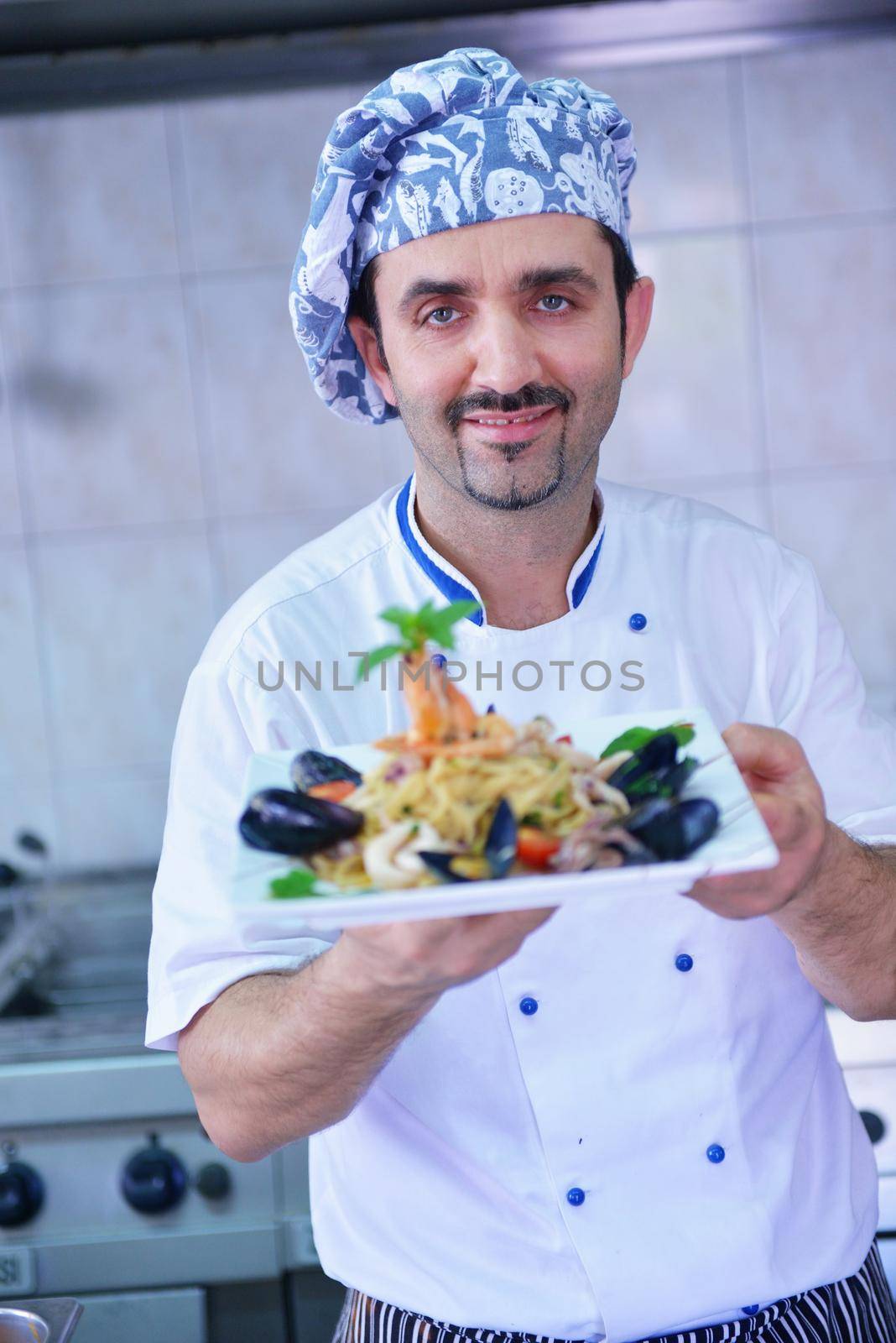 Handsome chef dressed in white uniform decorating pasta salad and seafood fish in modern kitchen